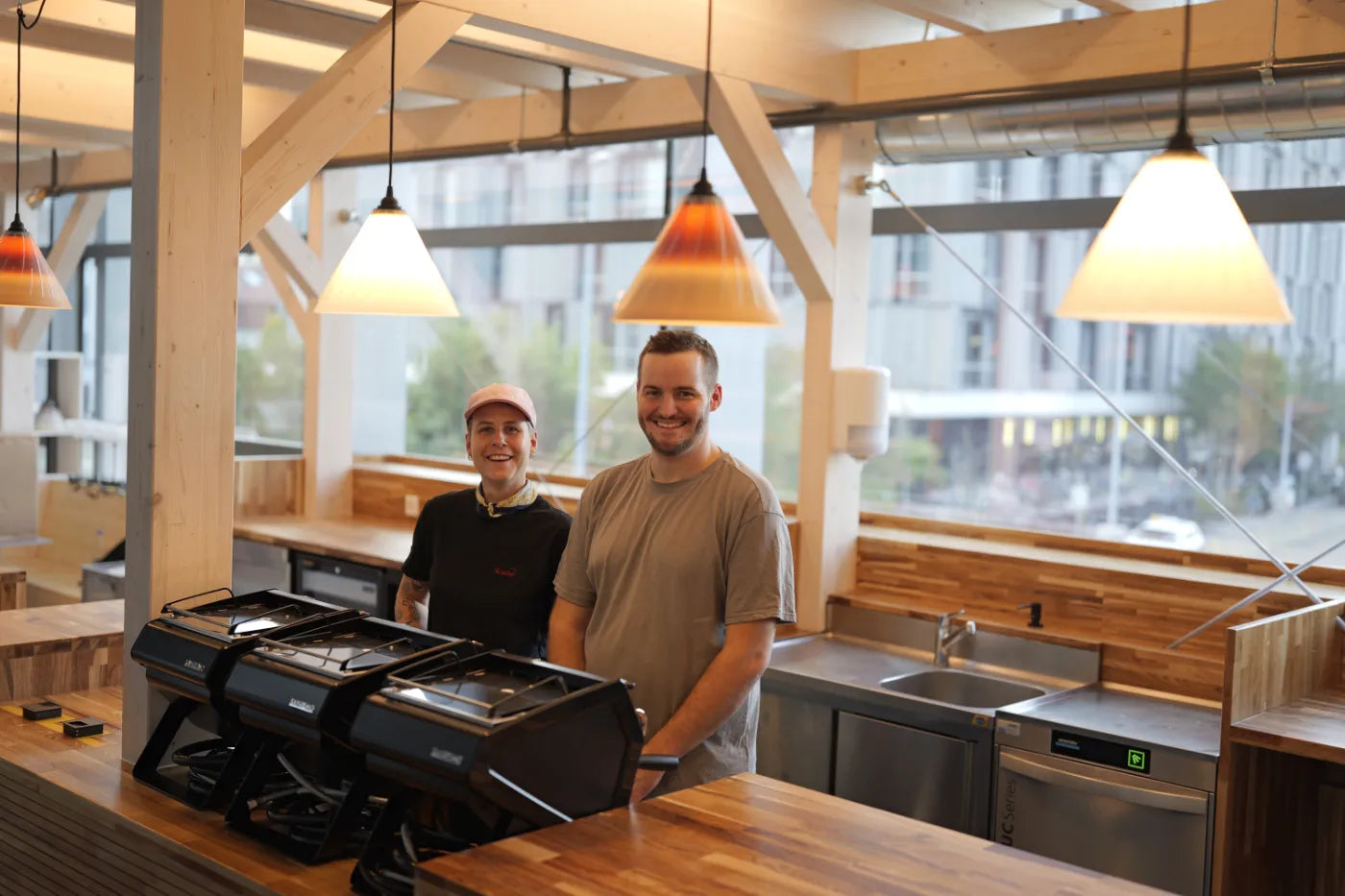 Interior of a modern cafe with two staff members behind a counter featuring an espresso machine, wooden finishes, pendant lighting, and large windows;