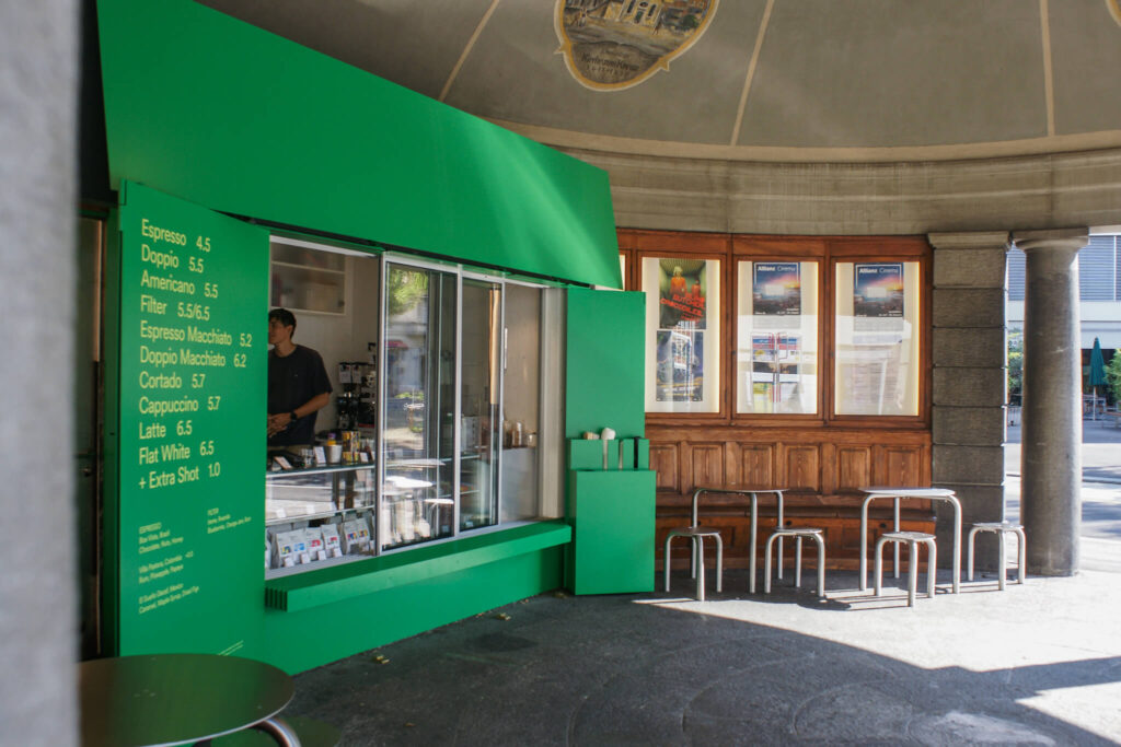 A bright green coffee kiosk with service window and menu board, housed in a historic pavilion with stone columns and domed ceiling