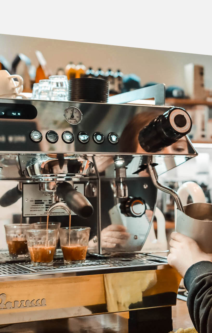 Close-up of a La Marzocco espresso machine in active use, with two shots pulling into glasses and a barista steaming milk