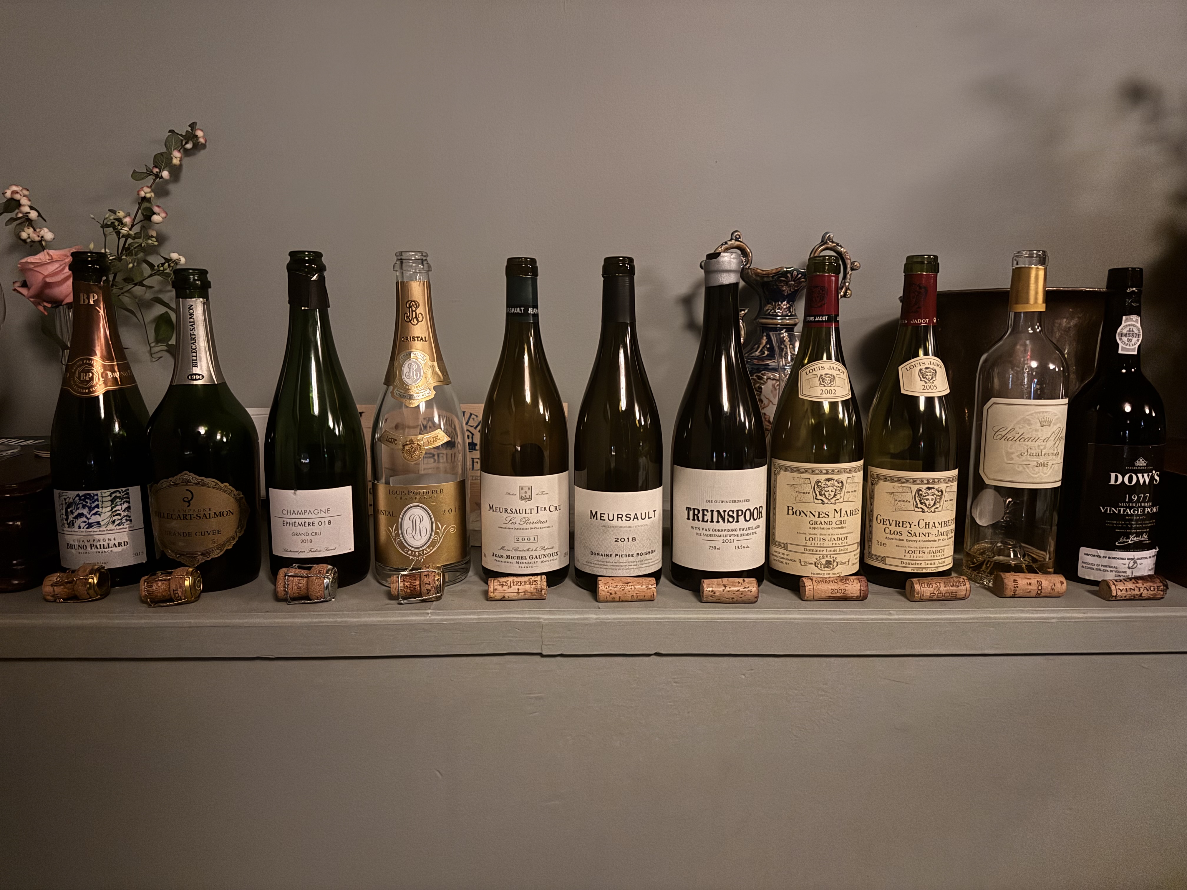 Wine bottles lined up on a shelf including Champagne, Meursault, Treinspoor, and Bonnes Mares with corks displayed below.