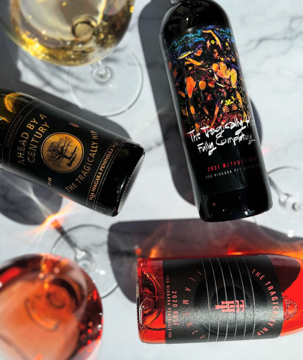 Two wine bottles on marble surface with wine glasses: Harlan Estate 2021 Cabernet and The Tragically Hip Complete 2021 Reserve VQA Niagara.