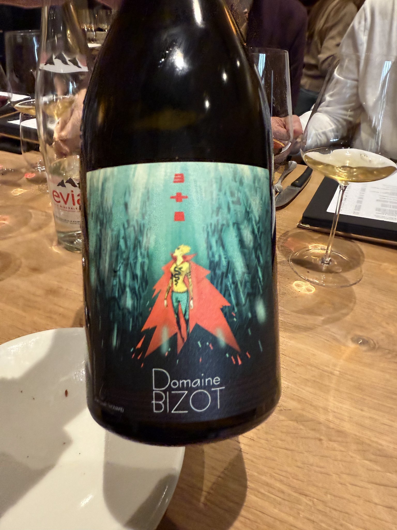 Domaine Bizot wine bottle with illustration of figure in red cape against teal background on wooden table