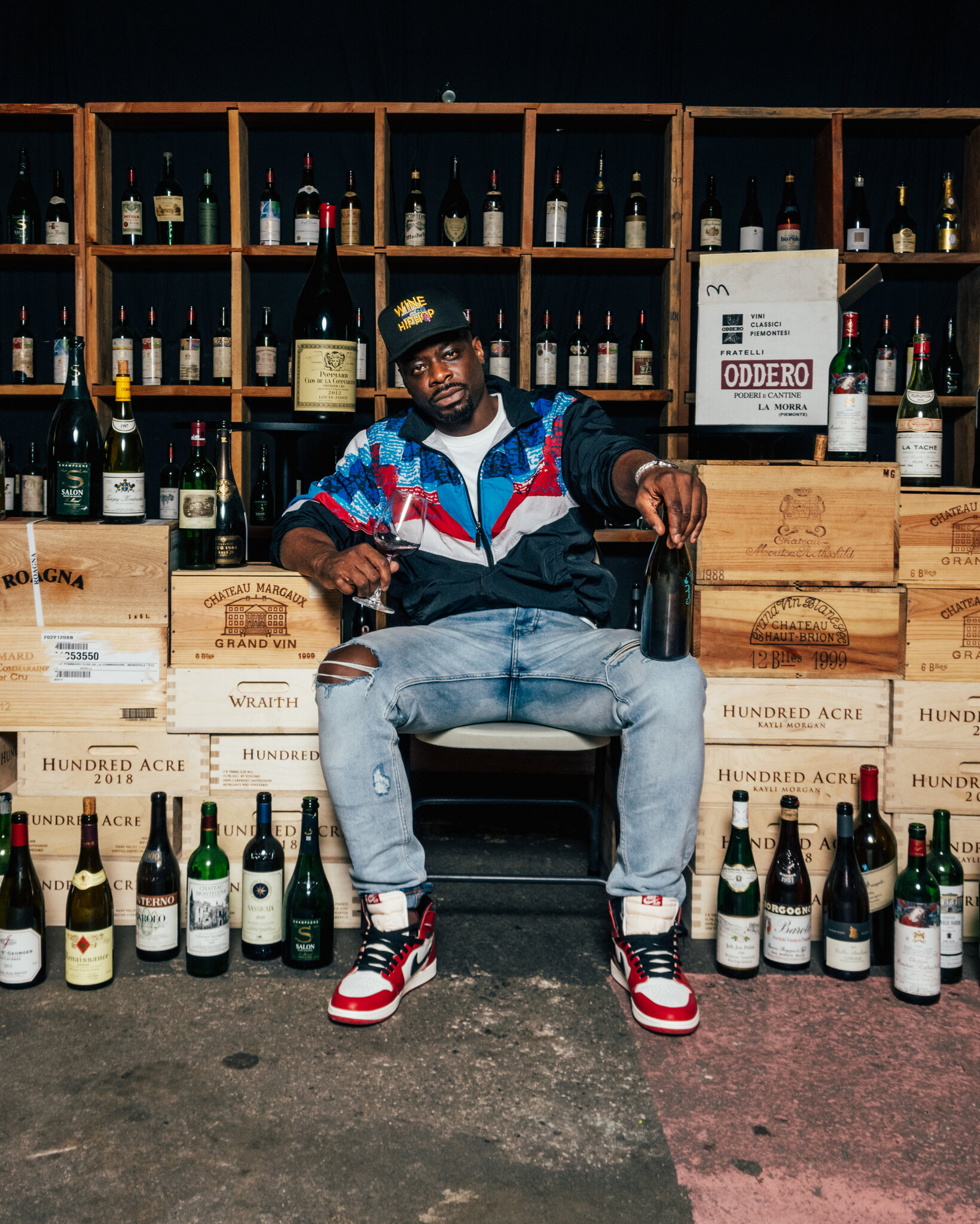 Man in black and blue jacket holding wine bottle seated on wooden wine crates labeled Château Margaux, Hundred Acre, and Wraith, with shelves of Odder