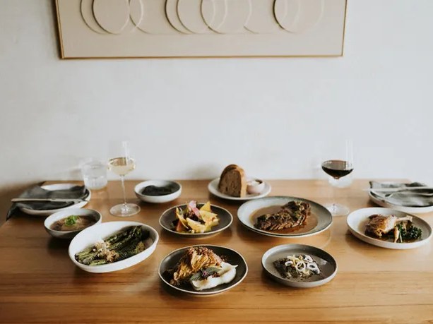 A wooden table is laden with an array of elegantly plated dishes, including roasted meats, asparagus, and various salads, alongside wine glasses and