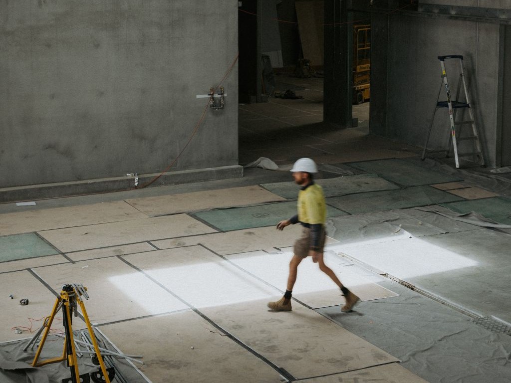 A construction worker in a hard hat and high-visibility vest walks across a large, tiled floor in an unfinished building, with concrete walls, a