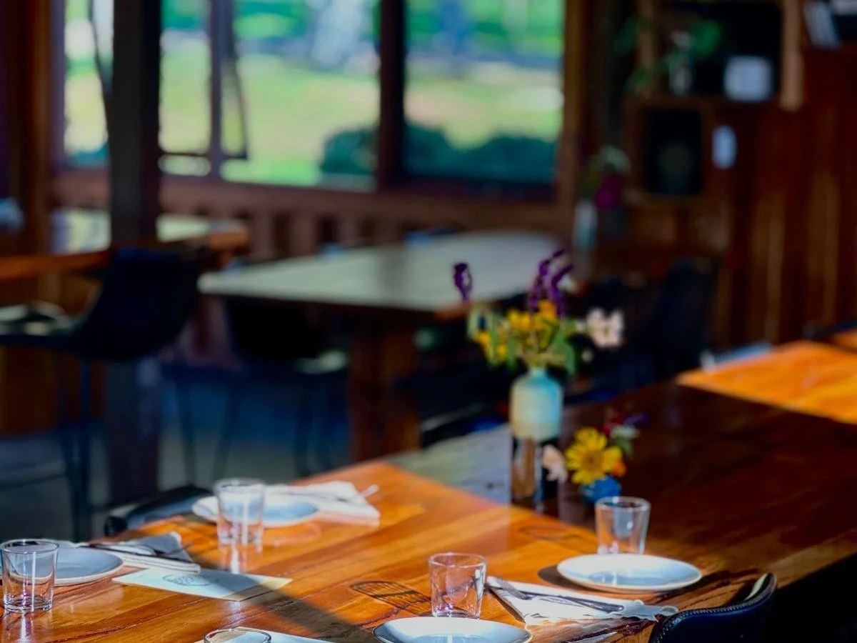 A warmly lit farm-to-table restaurant dining room interior featuring a long live-edge timber communal table set with white plates, glassware, and