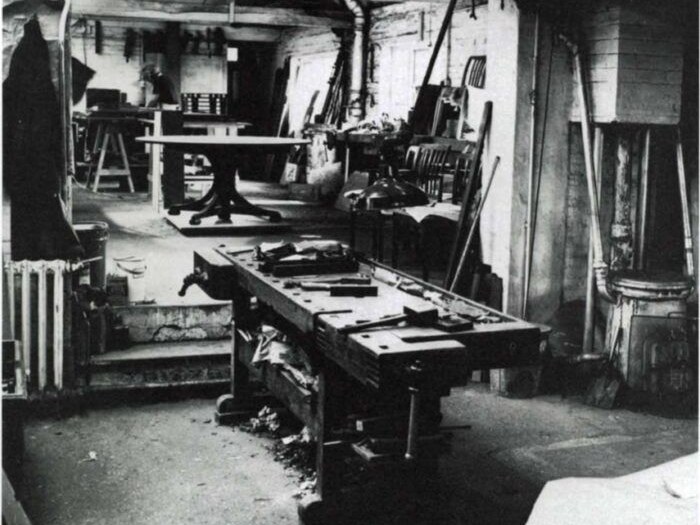 A black-and-white photo of the Edward Barnsley Workshop bench room, showing a workbench with tools and wood shavings, and furniture in the background.