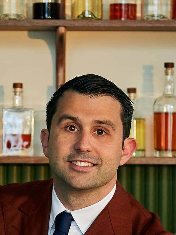 A professional portrait of Giorgio Bargiani, a man in a rust-colored suit, smiling behind a bar with empty cocktail glasses and bottles.