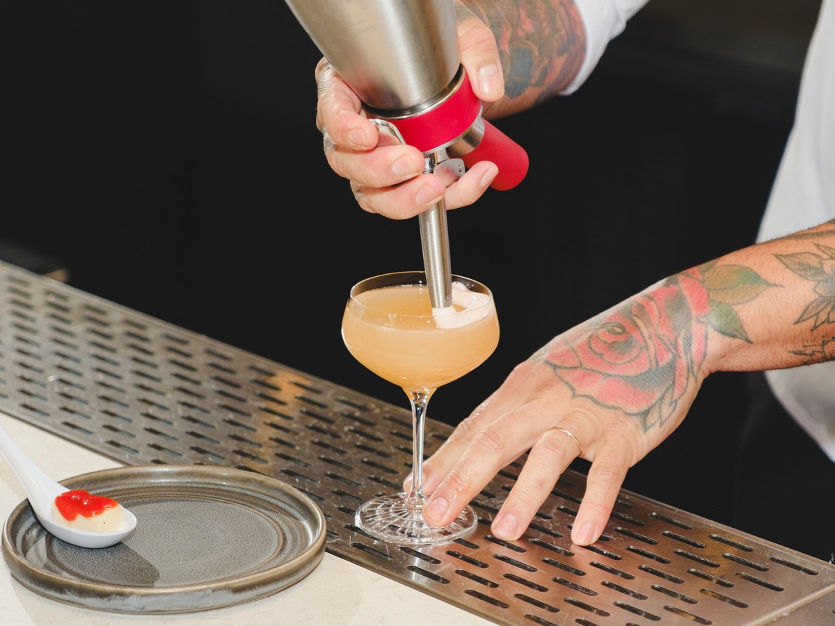 A close-up of a bartender's tattooed hands, one holding a metal cocktail dispenser pouring a creamy drink into a coupe glass, the other steadying the