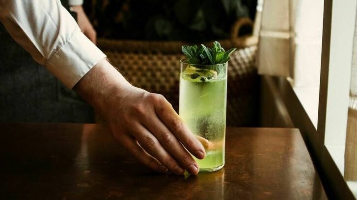 A bartender's hand in a white shirt cuff reaches towards a tall glass of green cocktail garnished with mint leaves on a wooden table.