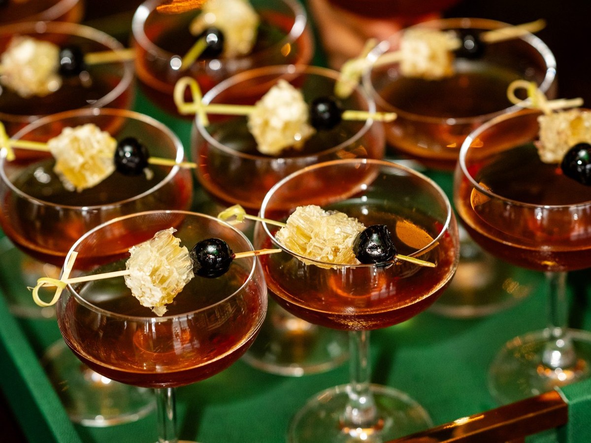 Multiple coupe glasses filled with amber cocktails, each garnished with a piece of honeycomb and a black cherry on a decorative pick, arranged on a