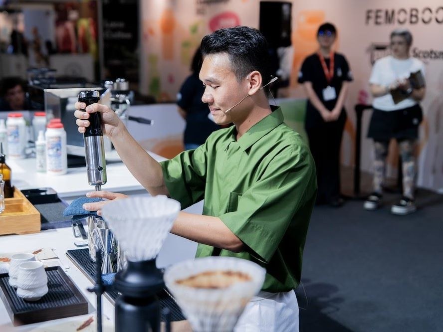 A barista in a green shirt performing a coffee demonstration at a professional event, with headset microphone and audience visible in background; sour