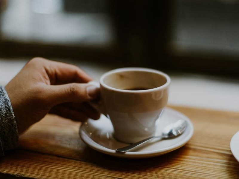 A person's hand holding a white ceramic coffee cup on a saucer with a spoon, placed on a wooden table.