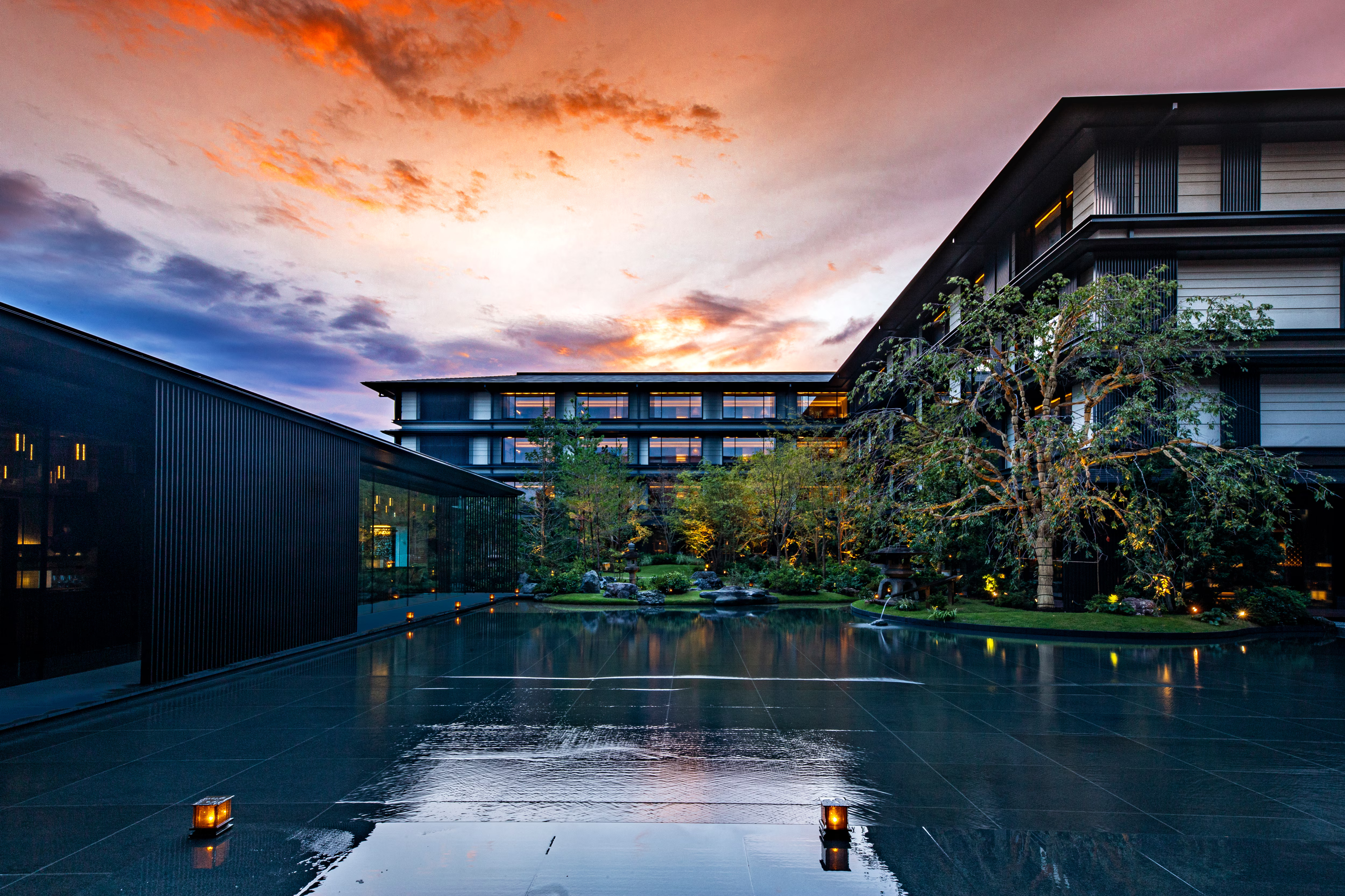 Modern building with glass walls at sunset, reflecting on a calm water surface. Trees and lanterns create a serene, peaceful atmosphere.