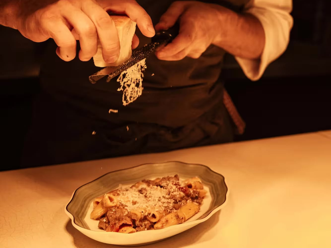 Chef grates cheese over Rigatoni alla Zozzona at Serra at The Chancery Rosewood under warm lighting in a rustic kitchen. Beige apron, white shirt, focused expression.