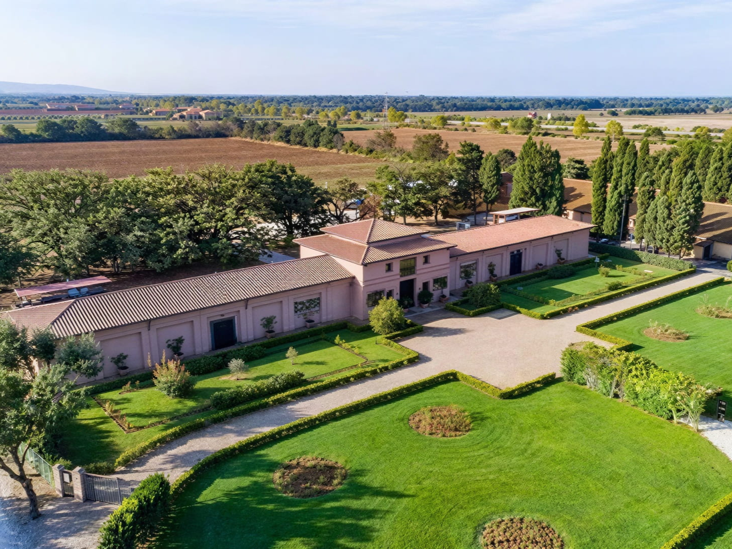 Aerial view of the new Tenuta San Gudio estate with a pink-roofed building, surrounded by manicured lawns, trees, and fields under a clear blue sky.
