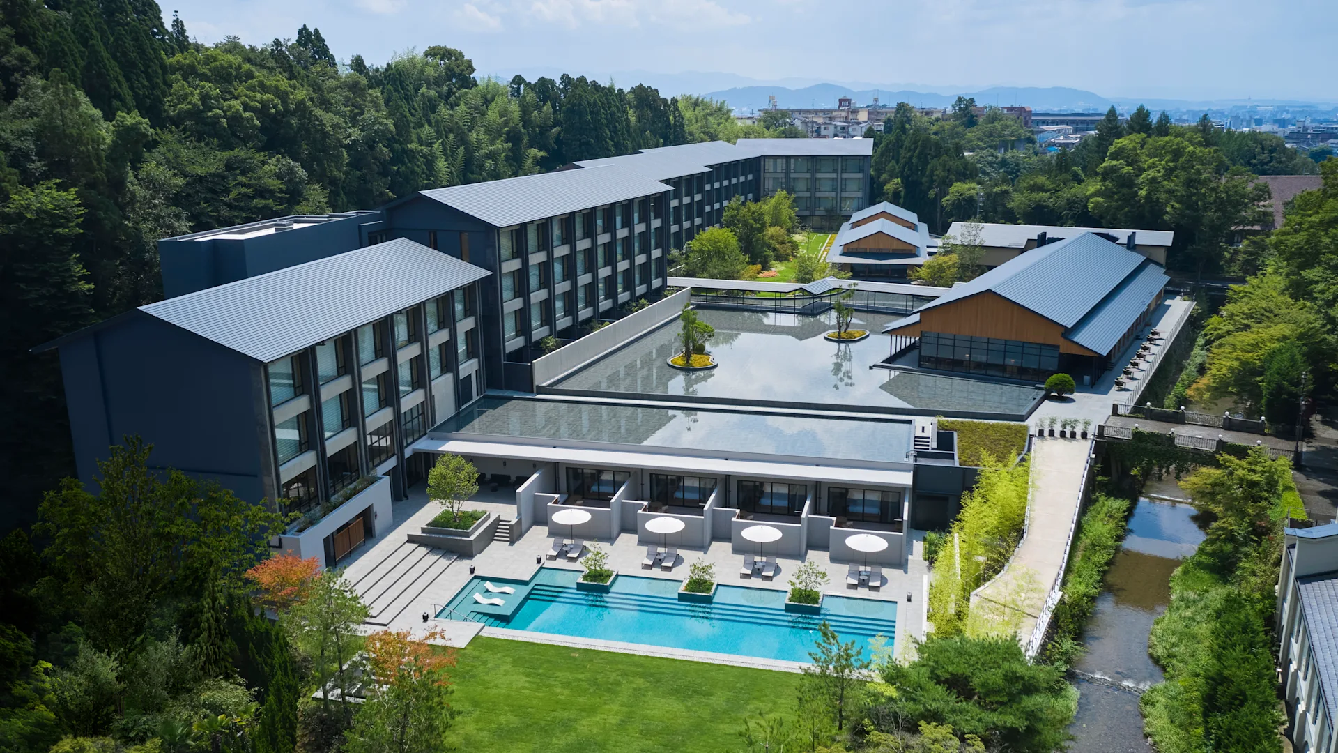 Aerial view of a modern hotel with a blue rooftop, courtyard pool, and lush green surroundings. Trees and cityscape background. Calm, sunny day.