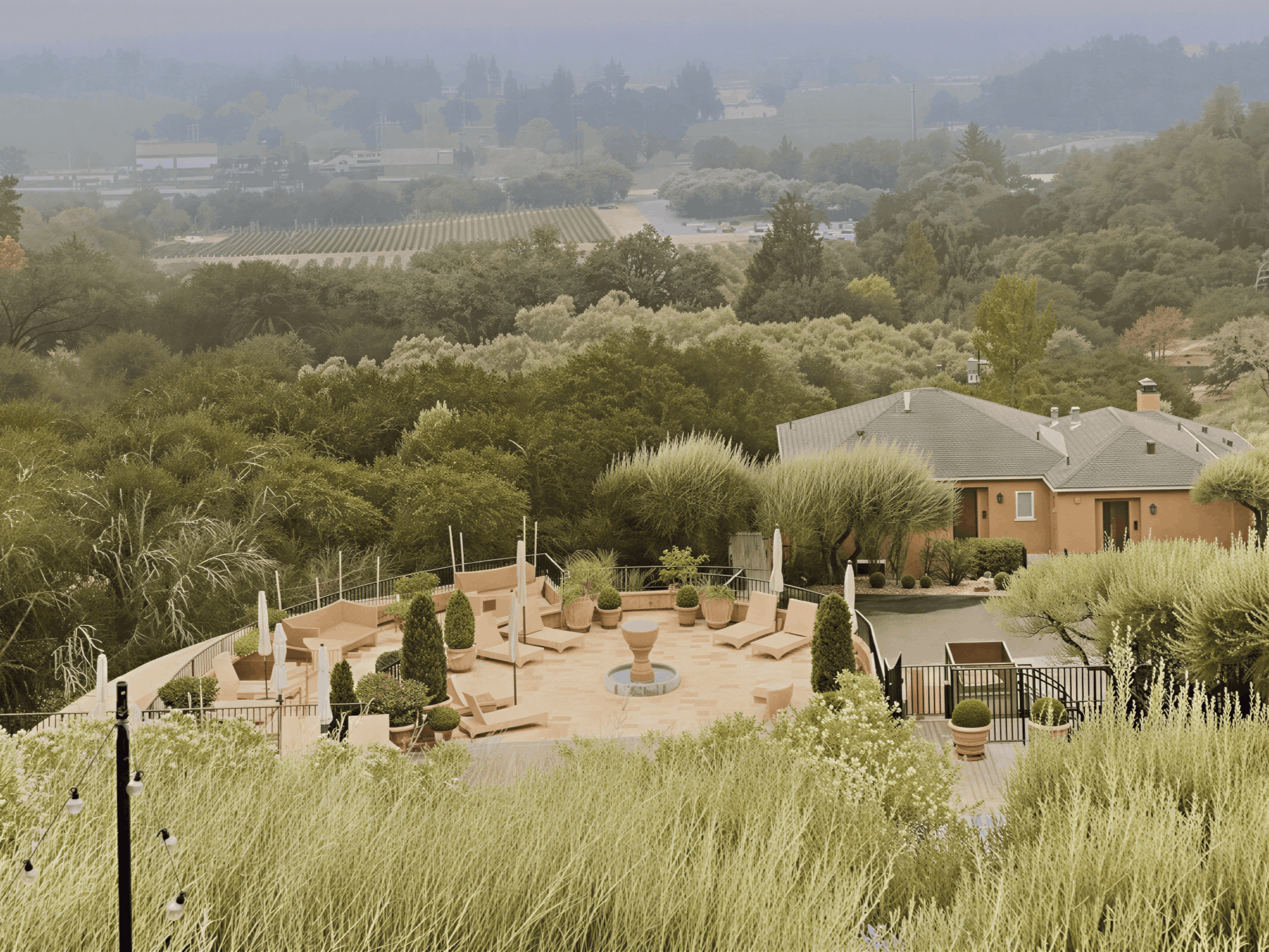 A patio with potted plants, lounge chairs, and a fountain overlooks a lush, green landscape. There's a building nearby and a misty backdrop.