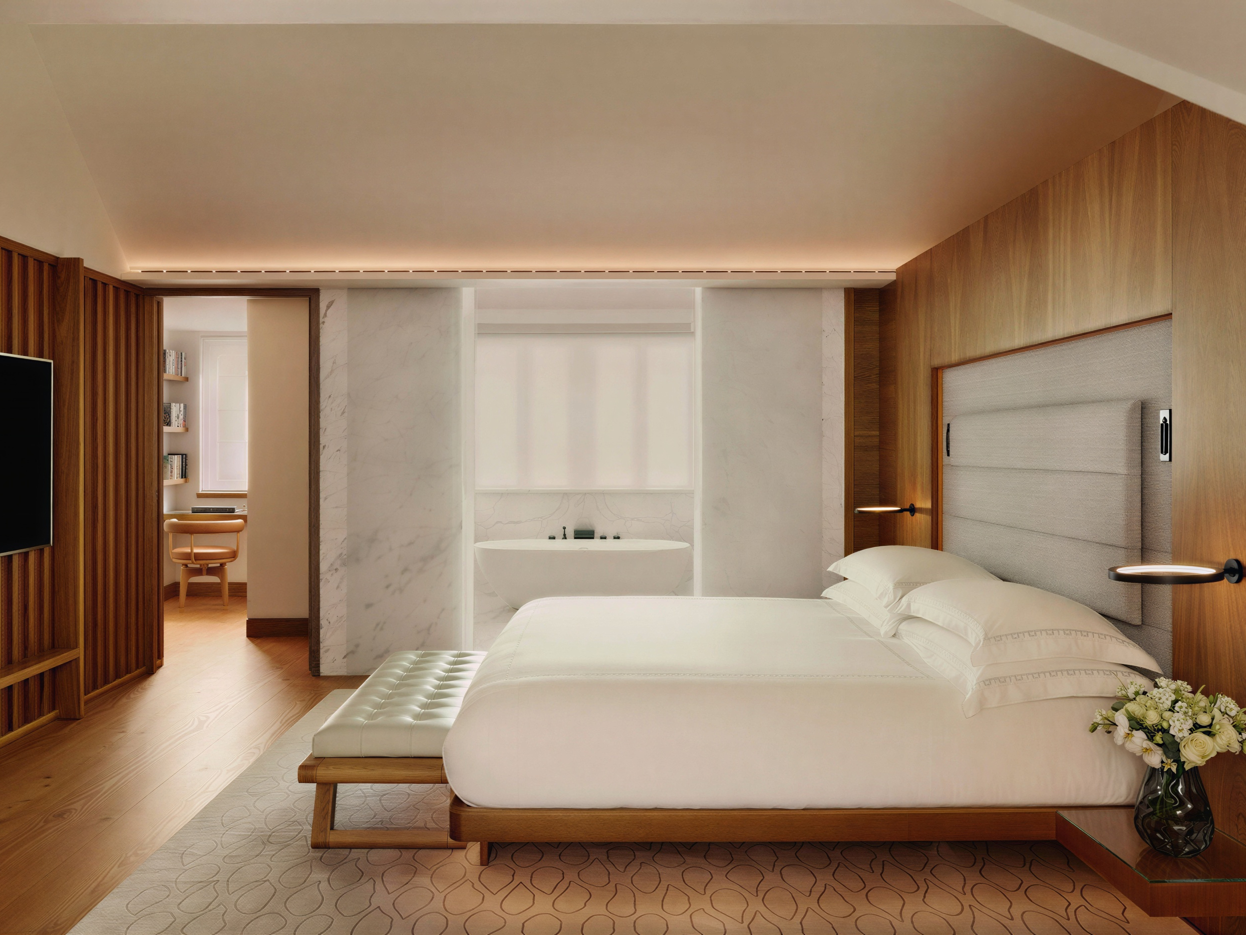 Modern bedroom with a white bed, wood accents, and marble walls. A vase of flowers sits on the bedside table. Calm, minimalist vibe.