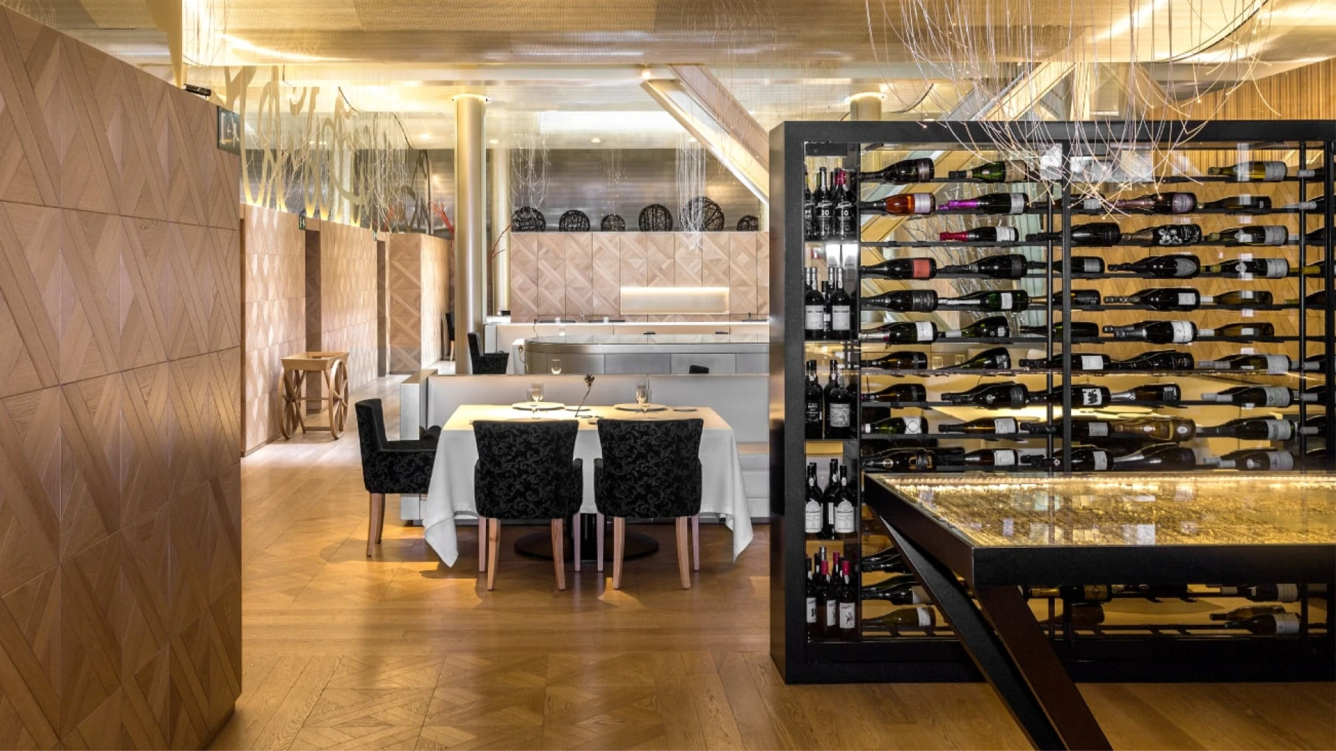 Elegant restaurant with wooden walls and floor, a wine display in the background, and a table set for two with black chairs and white tablecloth.