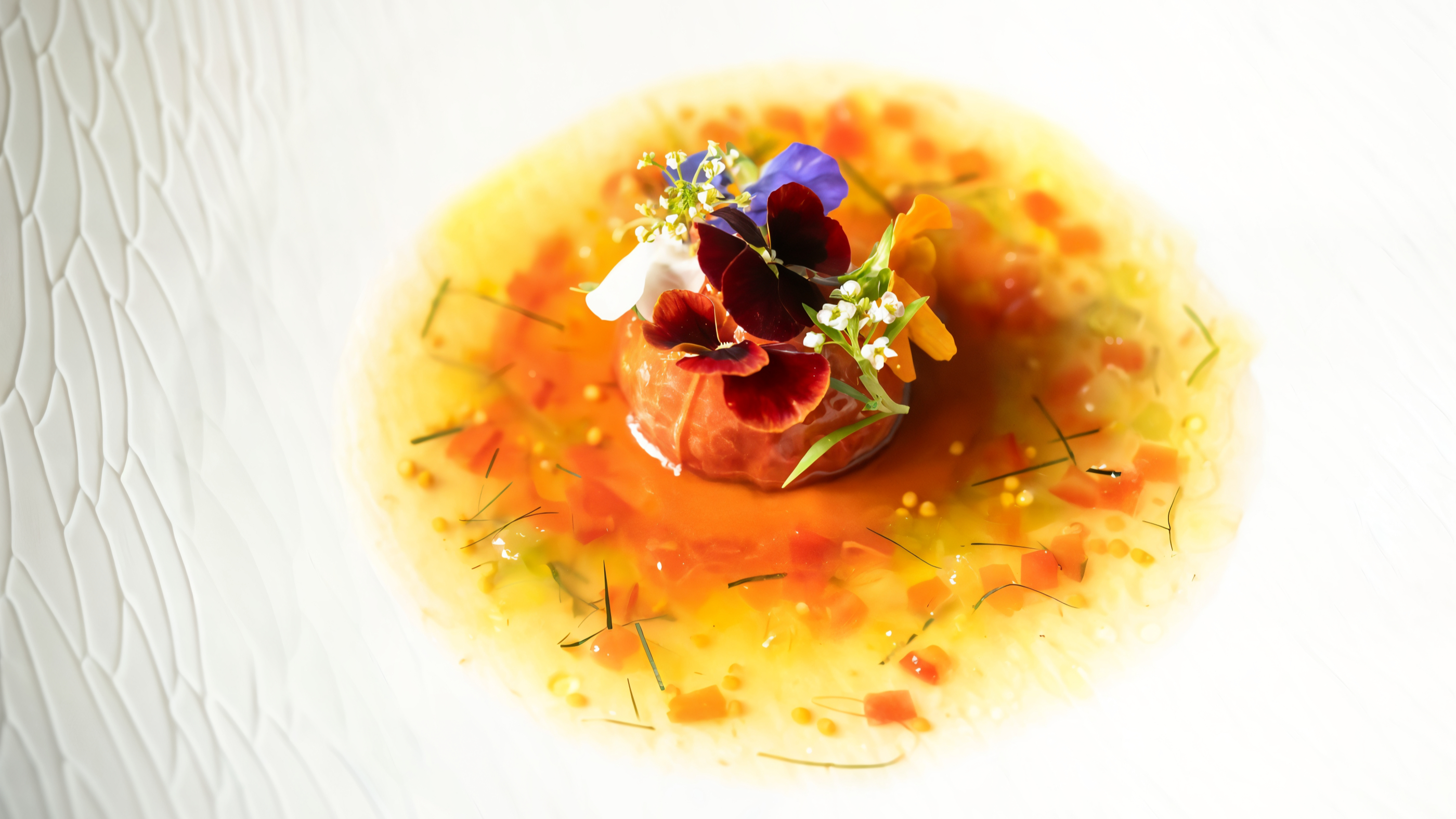 Gourmet dish with vibrant edible flowers on a tomato, surrounded by orange sauce, on a textured white plate. Fresh, colorful presentation.