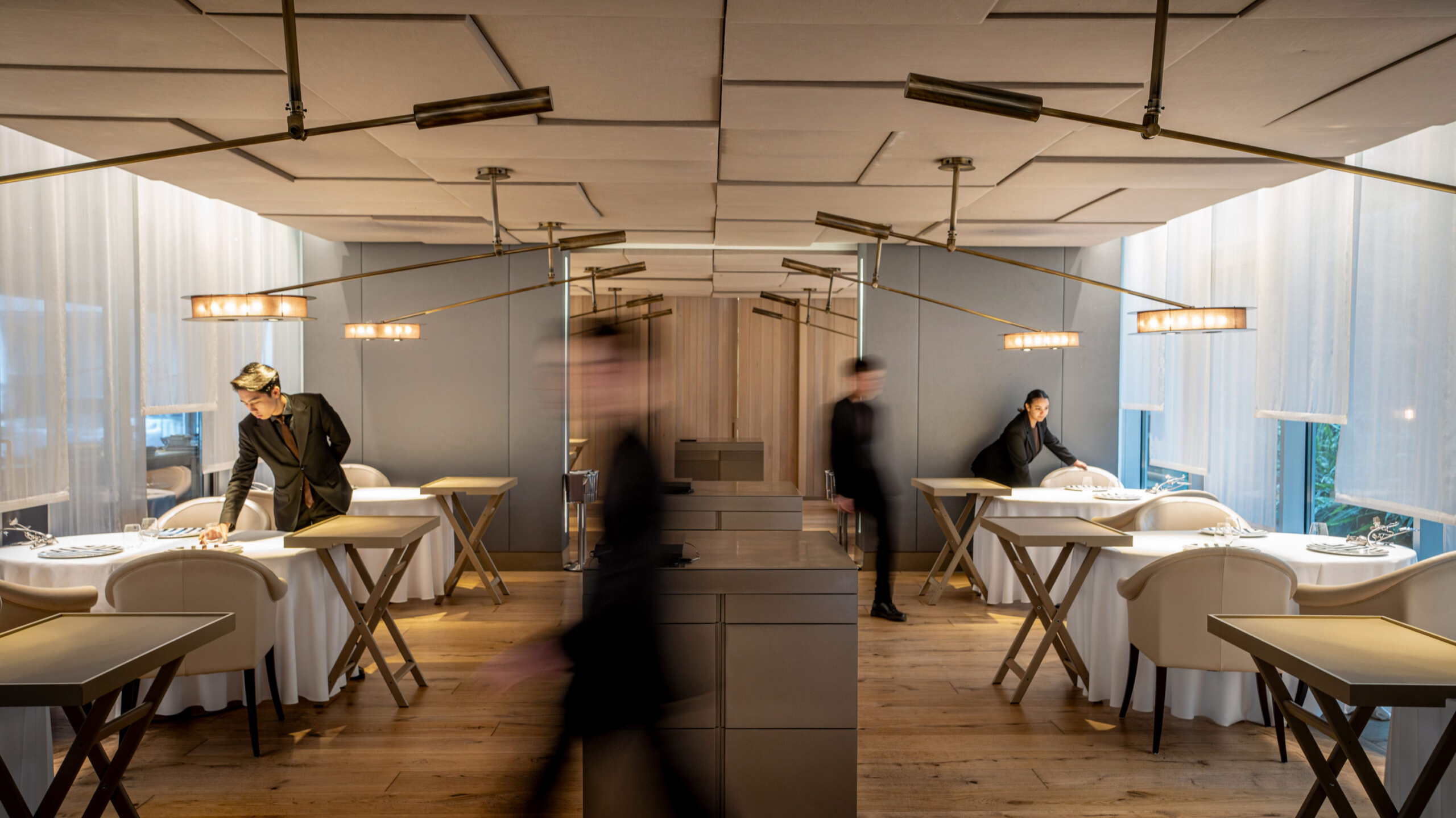 Restaurant scene with servers setting tables in a sleek, modern space. Soft lighting, wooden floors, and neutral tones create a serene atmosphere.