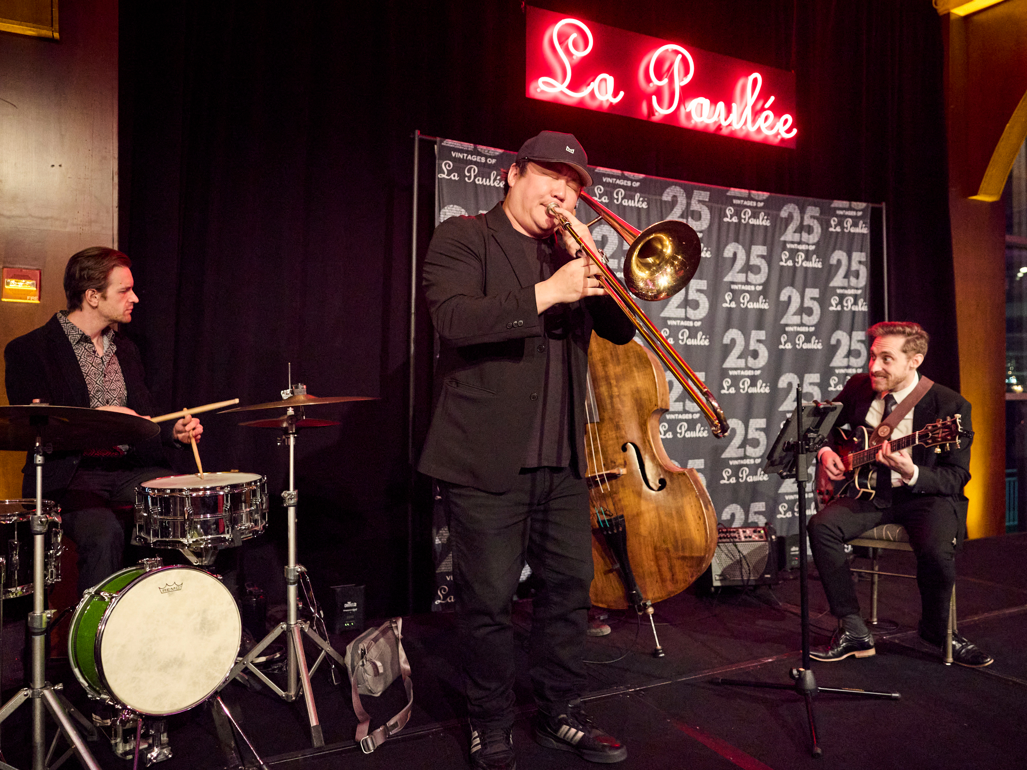 Jazz trio band plays at the Gala Dinner at La Paulée NYC 2025 performs in front of "La Paulée" neon sign; drummer, trombonist, and guitarist create lively ambiance on stage with black backdrop.