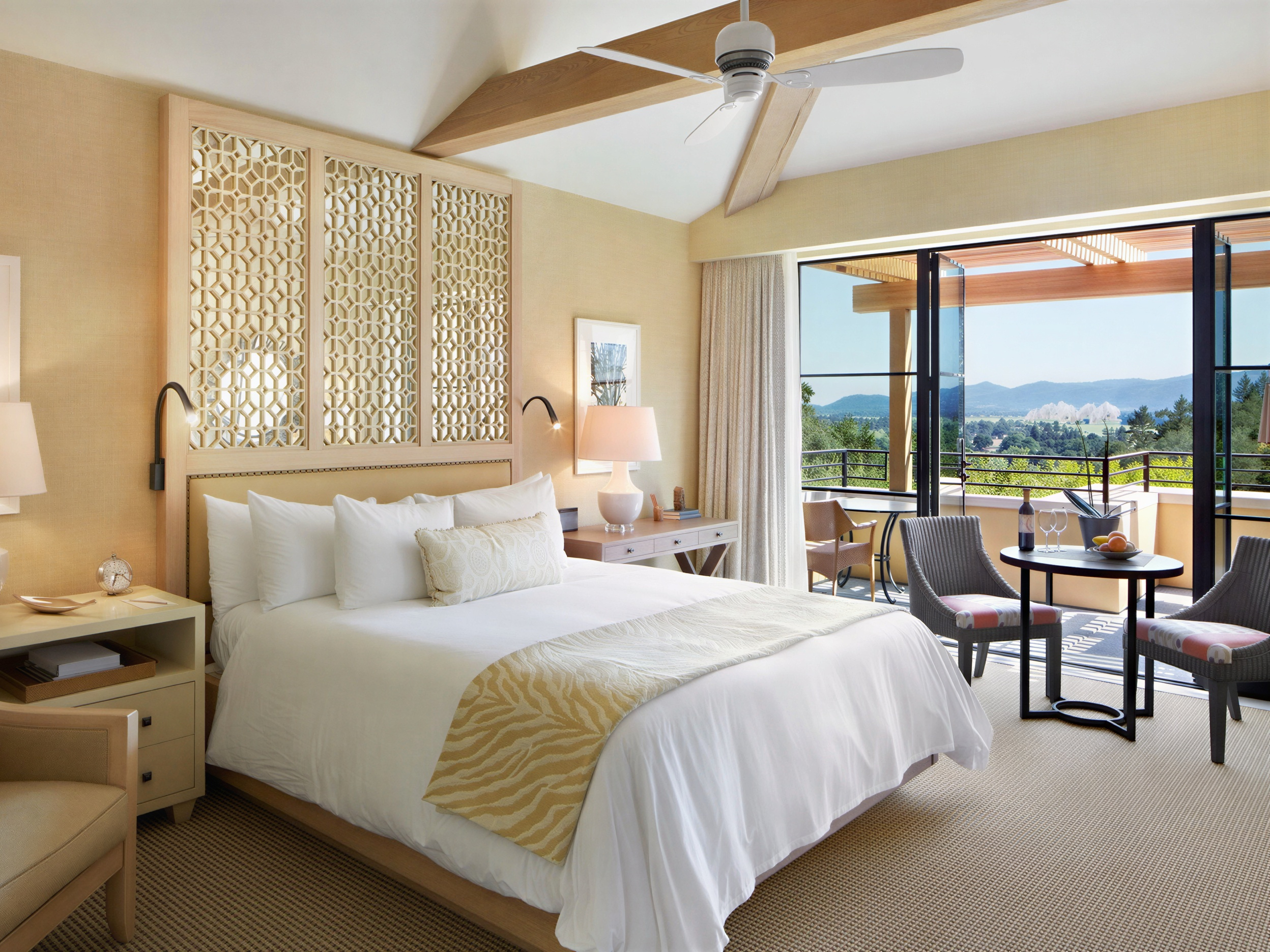 Bright bedroom with white bedding, decorative screen headboard, and balcony view of mountains. Table with wine, glasses, and fruit. Serene mood.