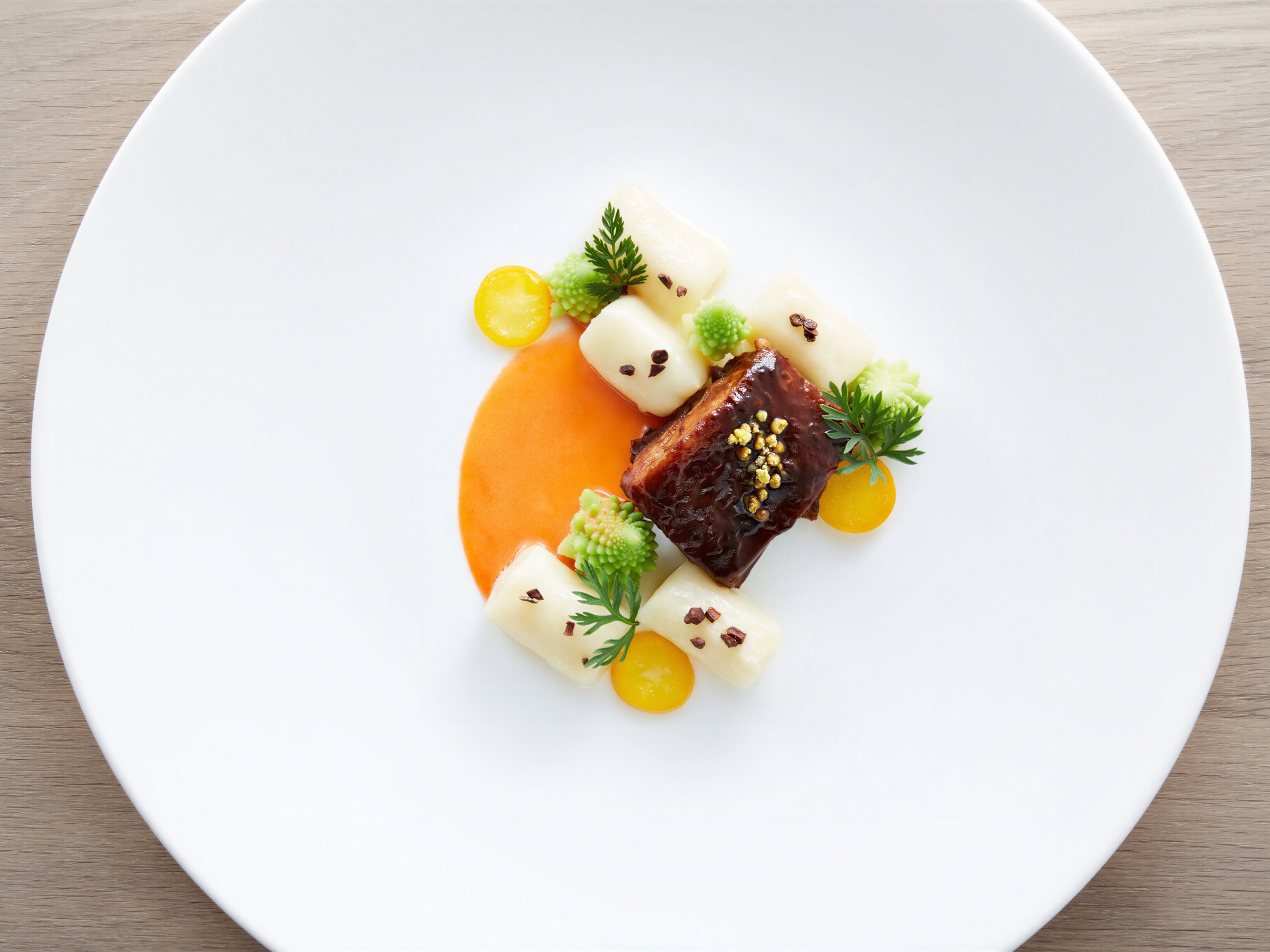 Gourmet dish on a white plate with glazed meat, creamy cubes, orange sauce, and green garnishes. Minimalist presentation on wood table.