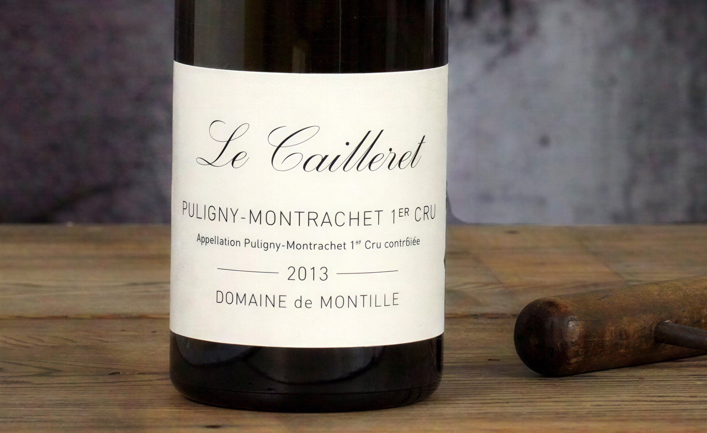 Wine bottle labeled "Le Cailleret Puligny-Montrachet 1er Cru 2013" on a wooden table with a rustic background.