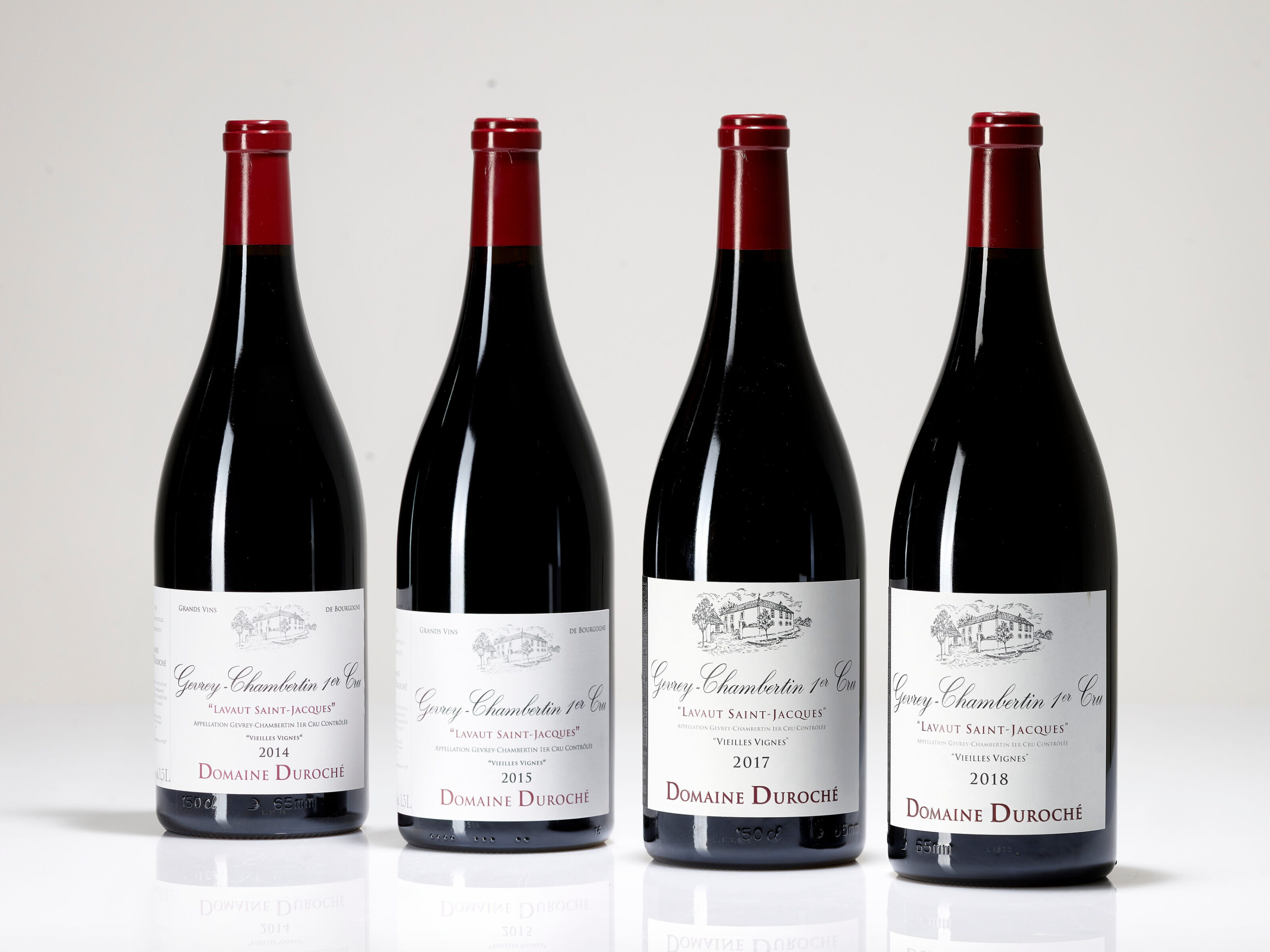 Four wine bottles of 2014, 2015, 2017 and 2018 Domaine Duroche Gevrey Chambertin Lavaut Saint-Jacqueswith red caps stand on a white surface. Labels display years 2014-2018, "Domaine Duroche." Simple white background.