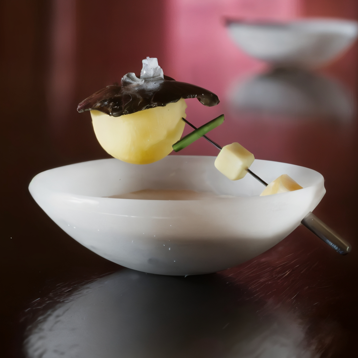 White bowl with speared cheese, apple slice, and mushroom on shiny surface. Pink background adds contrast to the minimalist setup.