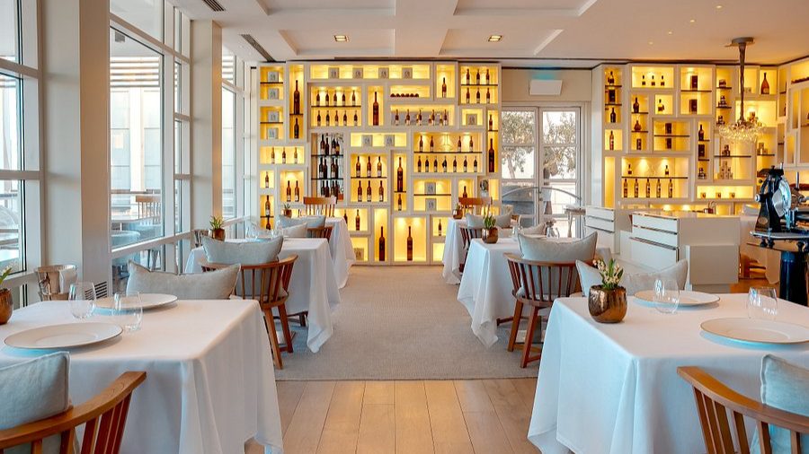 Elegant restaurant with white tablecloths, wood accents, and warm-lit shelves displaying bottles. Large windows create an inviting ambiance.
