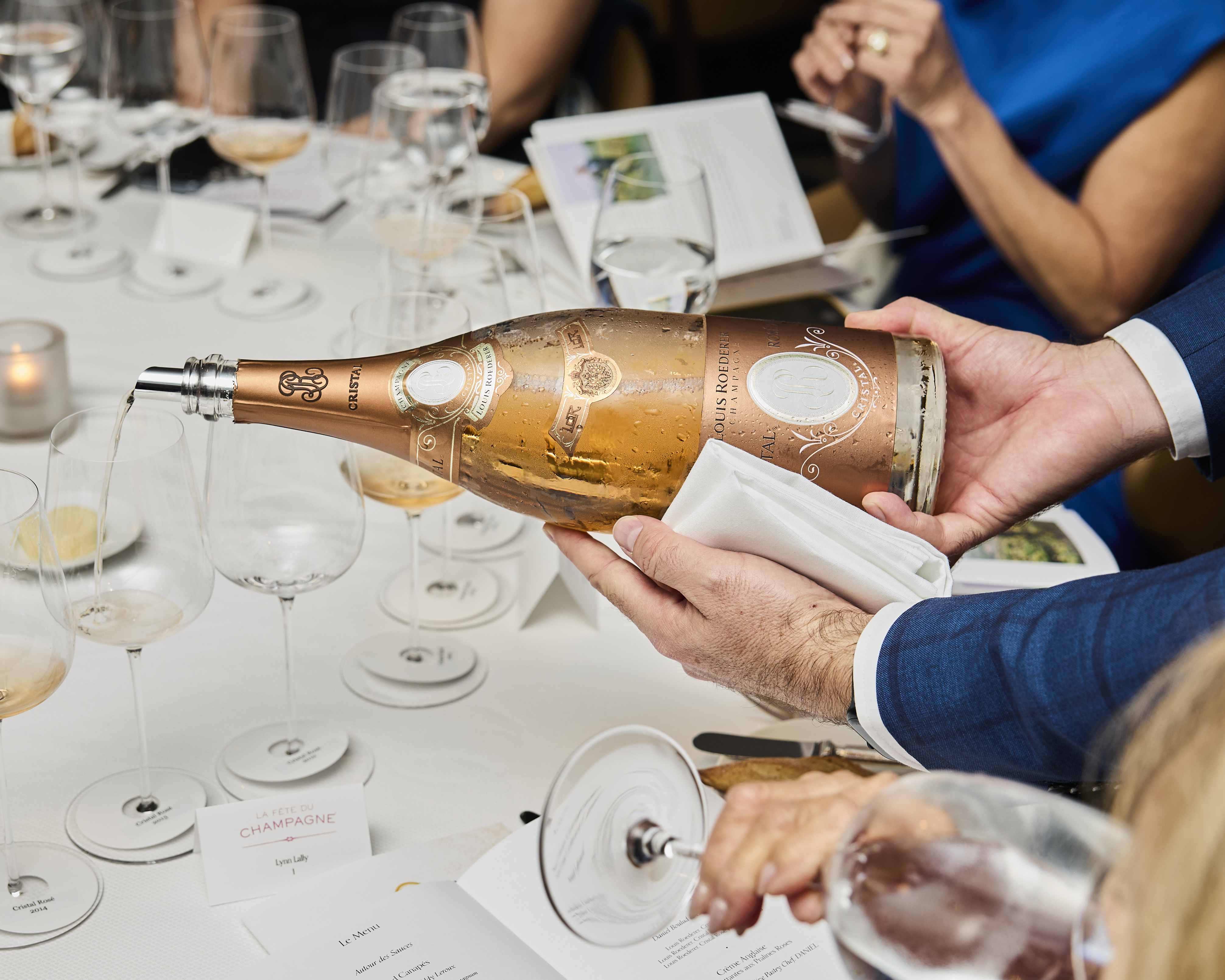 Hands in a blue suit pour Louis Roederer champagne from a large, bronze bottle into a glass on a white tablecloth with elegant setting.