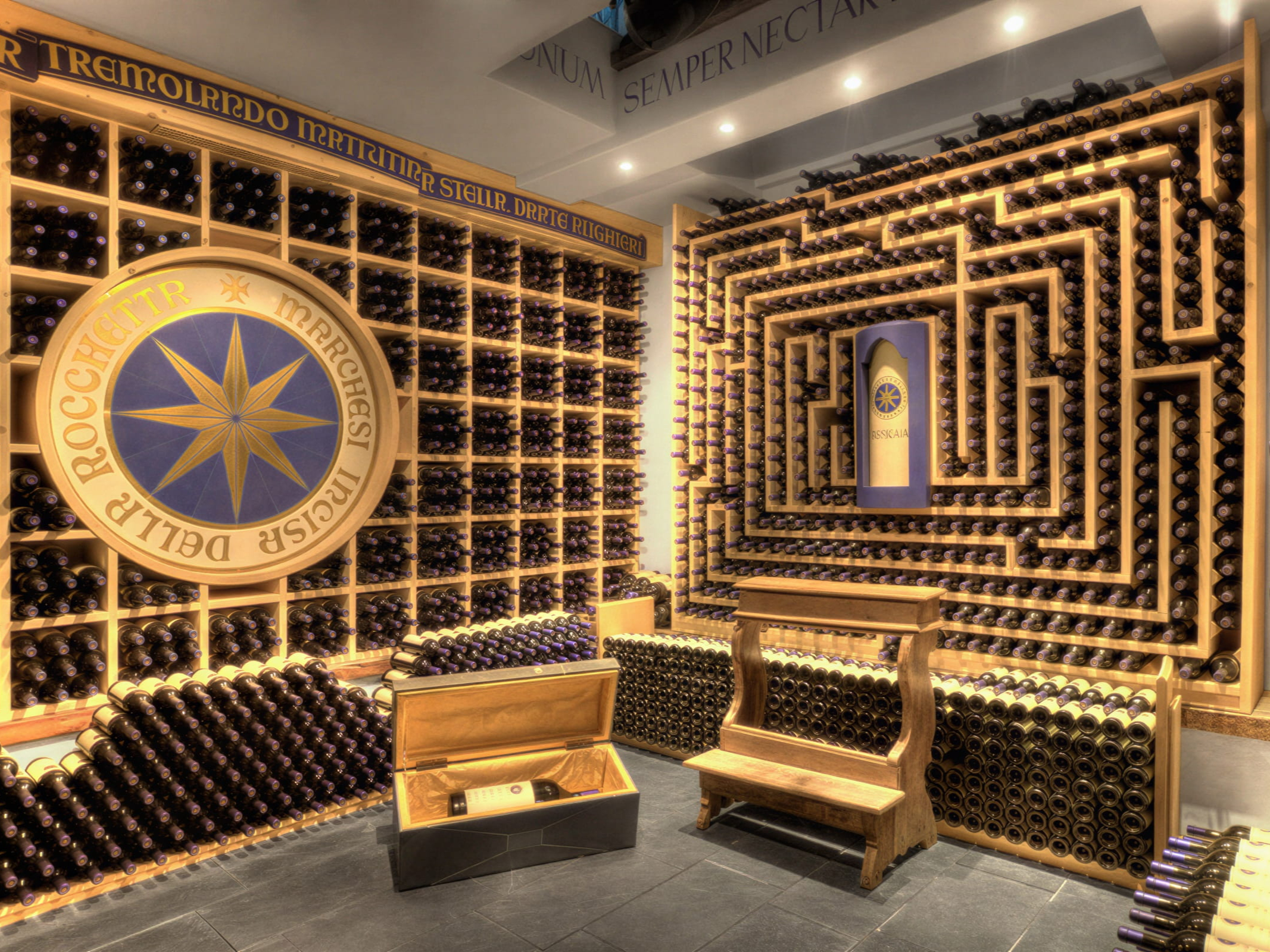 Historical wine cellar of Sassicaia at Tenuta San Guido with wooden racks filled with hundreds of wine bottles, a large compass rose emblem, and a wooden bench, creating an ornate display.