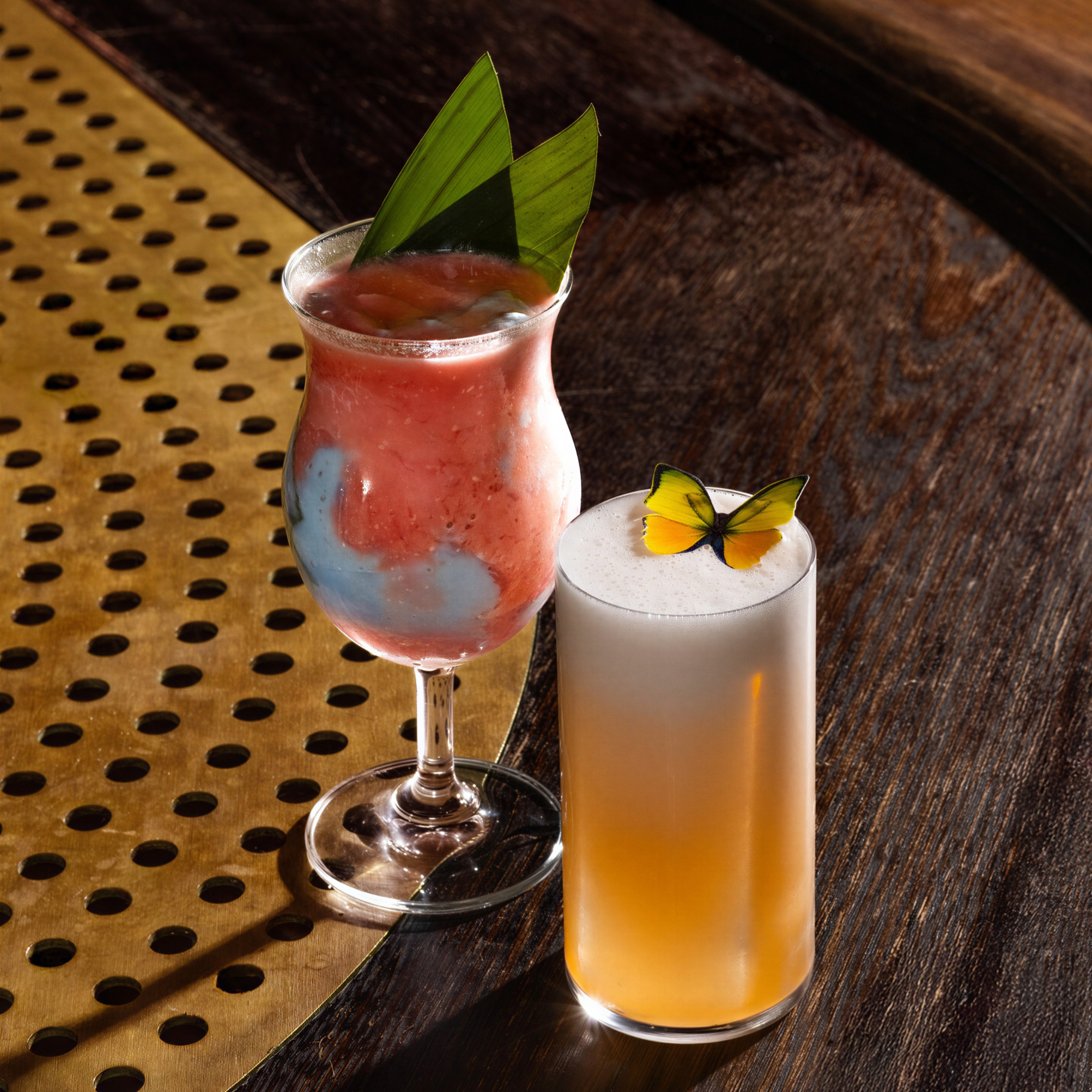 Two colorful cocktails on a wooden and brass table. One has a butterfly garnish, and the other is red and blue with green leaves.