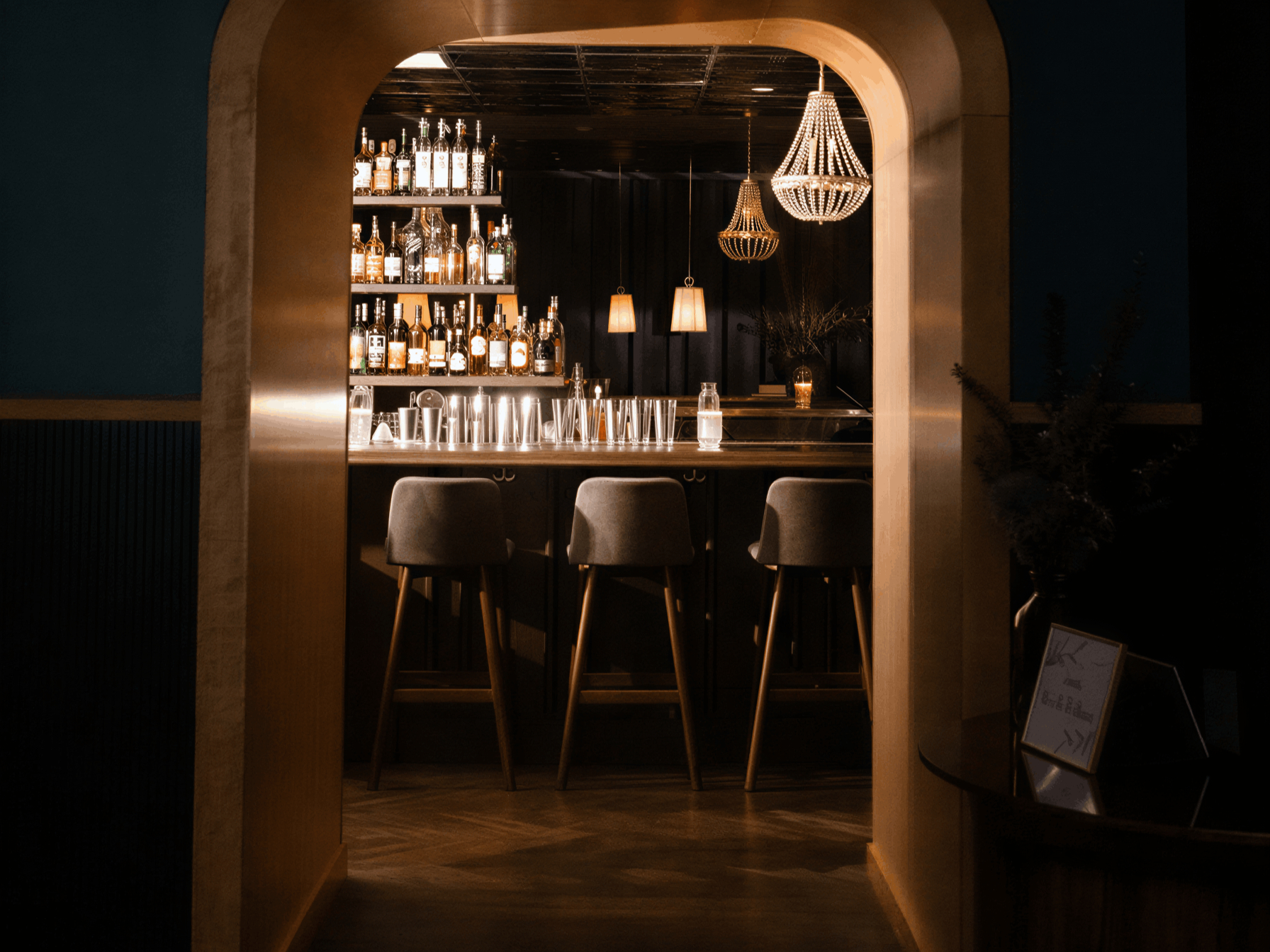 Dimly lit bar with a chic chandelier, liquor bottles on shelves, and three empty stools at the counter, evoking a cozy, elegant ambiance.