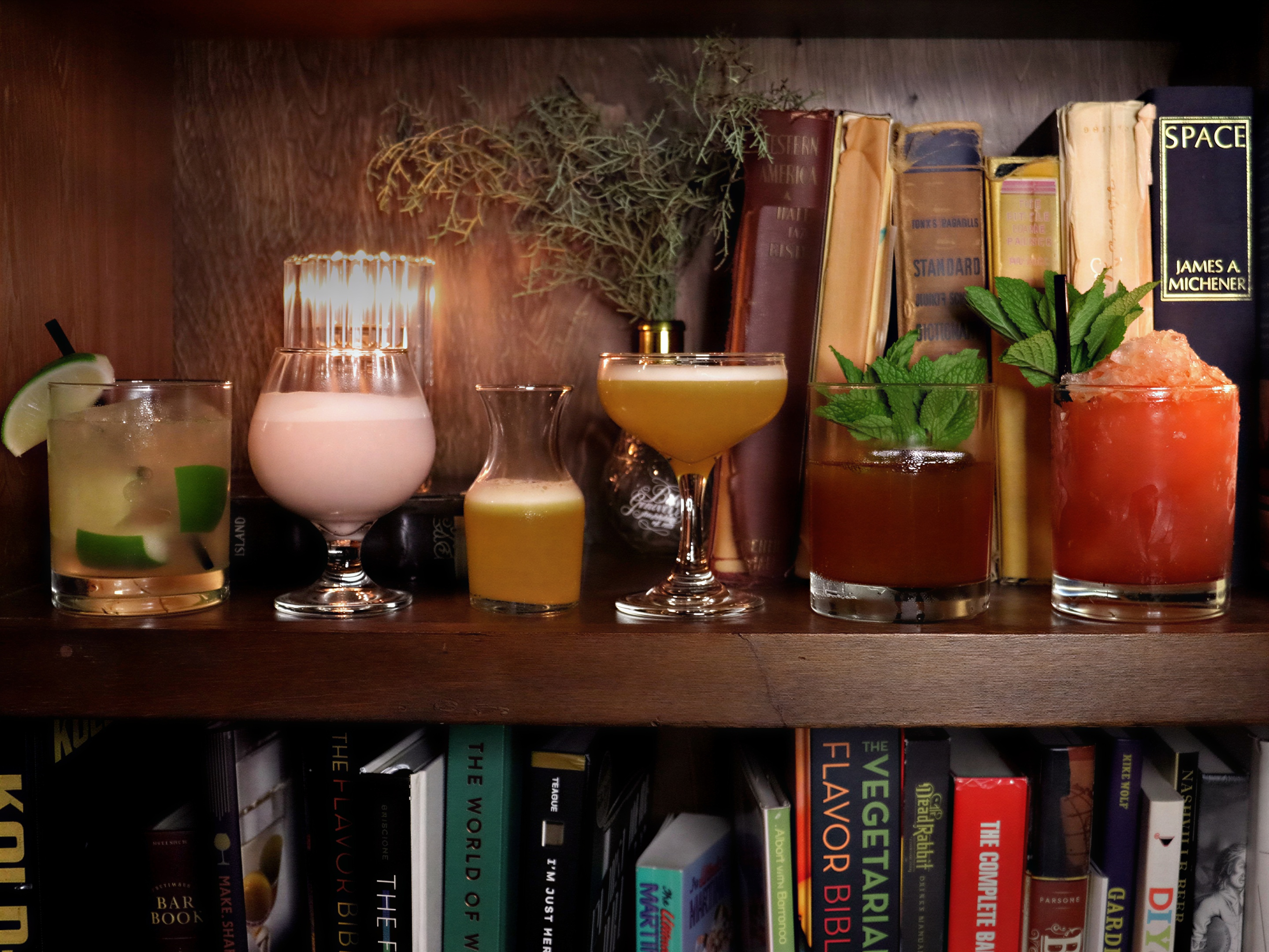 Assorted cocktails on a wooden shelf in front of books, featuring vibrant drinks with garnishes. The mood is cozy and inviting.
