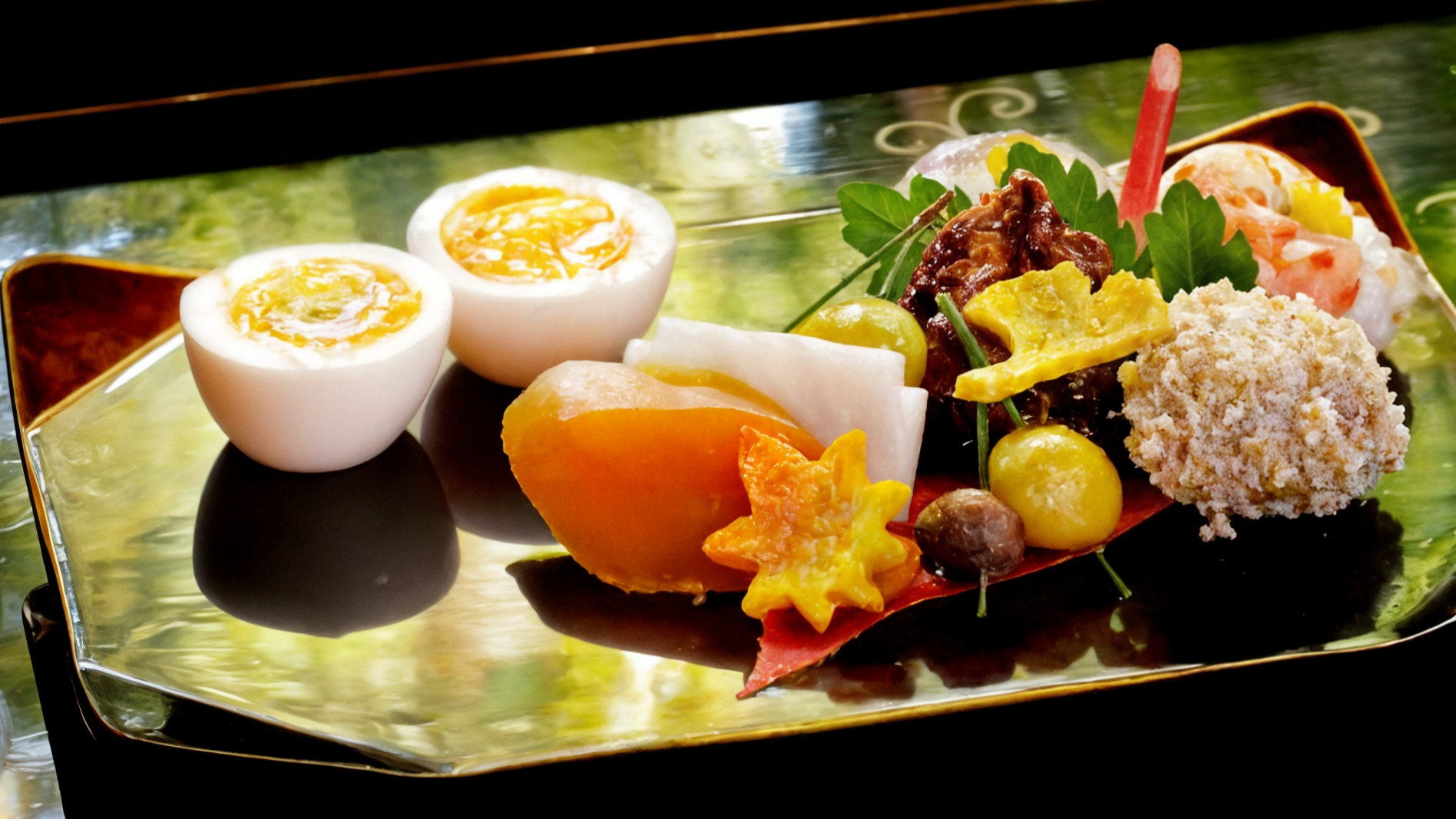 Elegant Japanese dish with halved eggs, orange star-shaped garnish, and assorted colorful foods on a reflective tray. Lush greenery background.