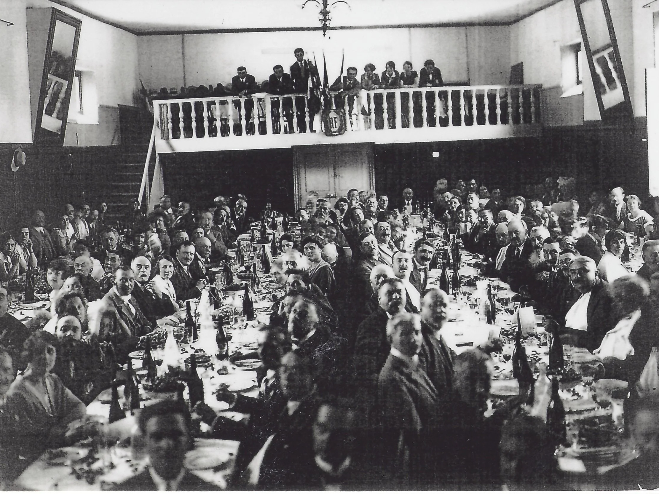 The historic La Paulée de Meursault dinner in Burgundy, France in a hall with high ceiling. Guests seated at long tables with bottles, chandeliers overhead, balcony visible.