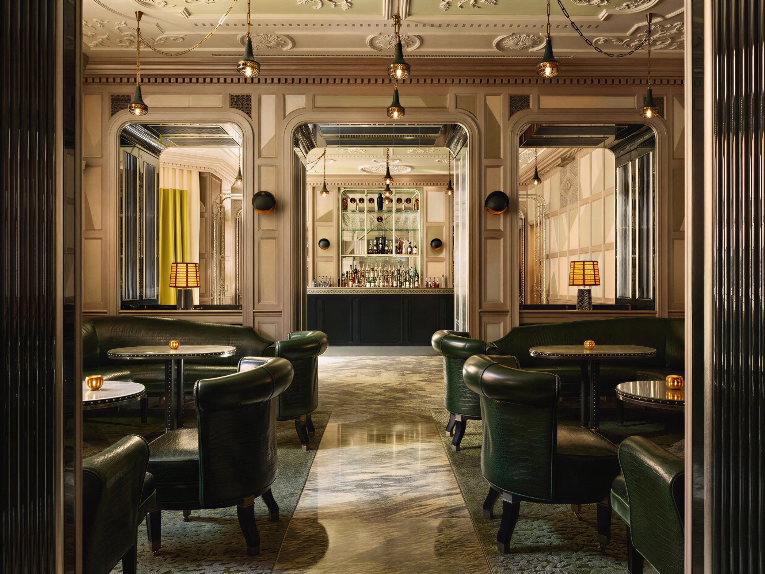 Elegant bar interior with dark green leather chairs, round tables with candles, arched mirrors, and a stocked bar. Warm lighting.