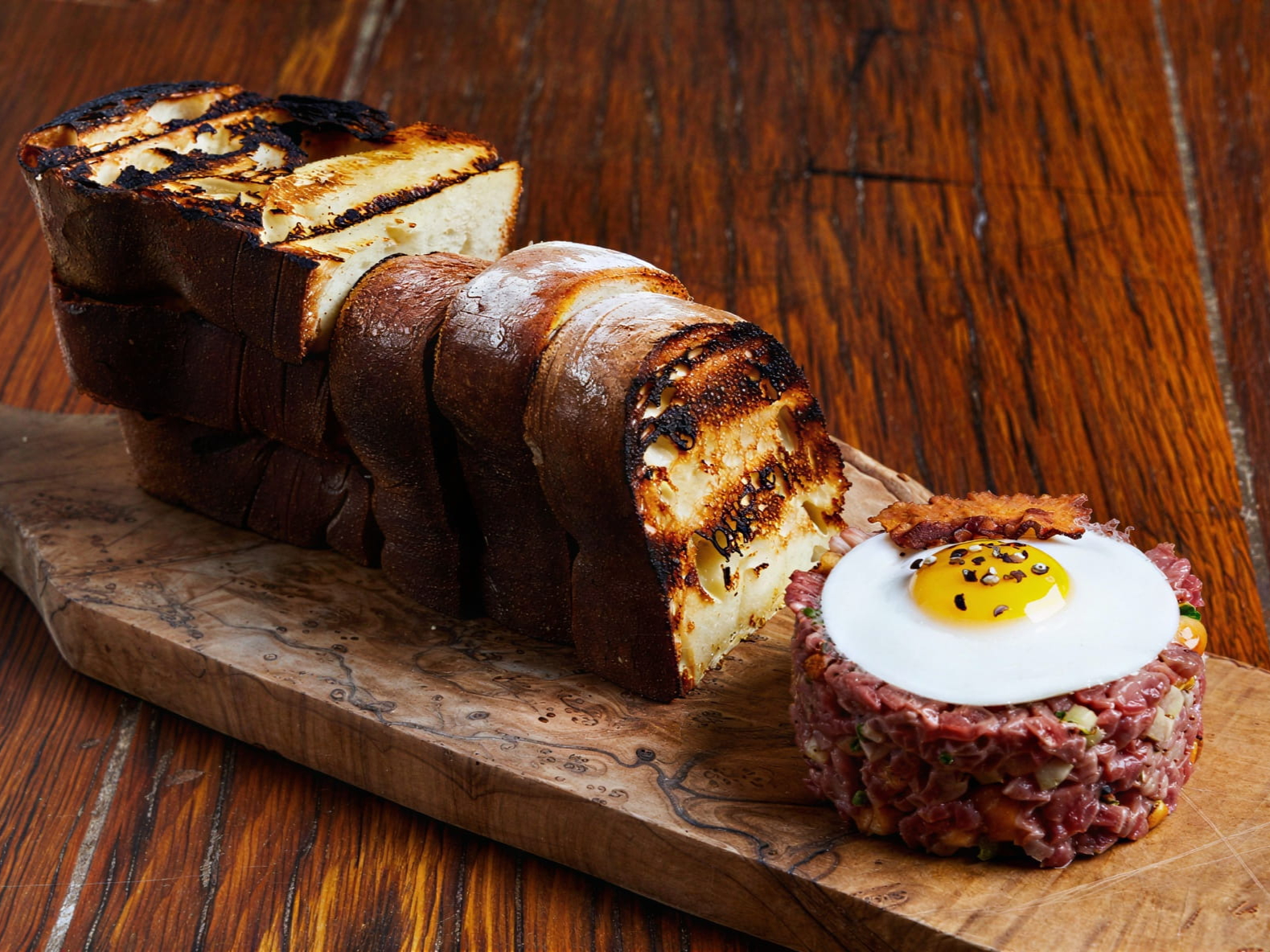 Grilled bread and Aged Wagyu Beef Steak Tartare Piemontese from Locanda Verde with a sunny-side-up egg and bacon on a wooden board, set against a rustic wooden background.