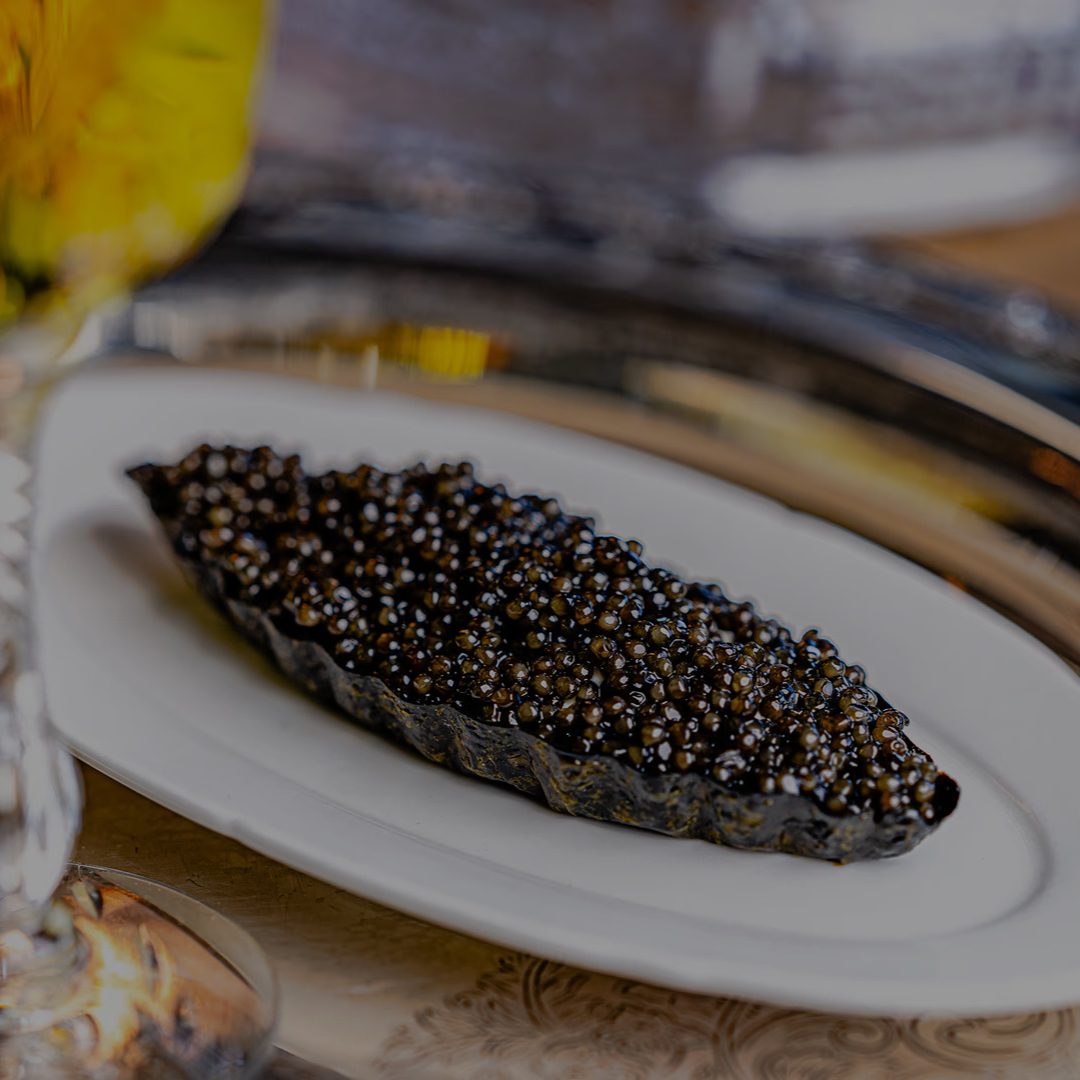Caviar on a decorative metal tray, served in a ceramic dish. Crystal glass with yellow-green drink nearby adds elegance. Rich, luxurious setting.