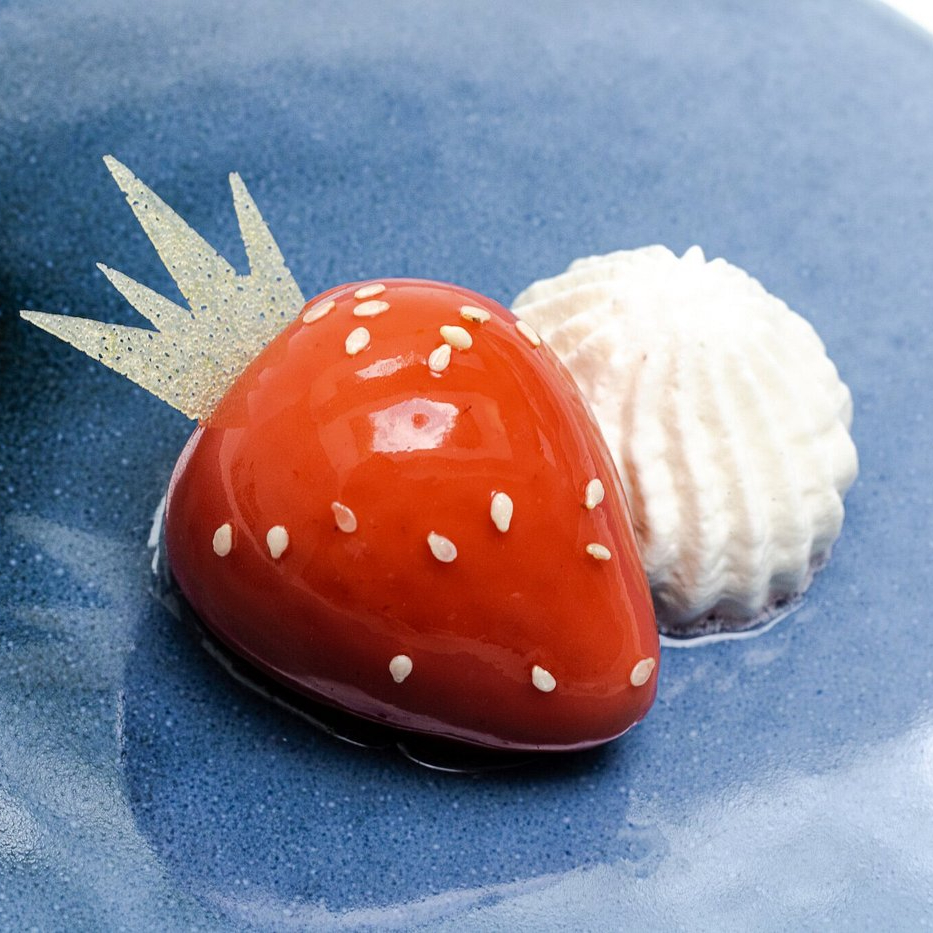 Shiny red strawberry-shaped dessert with sesame seeds and a white cream swirl on a blue plate. Elegant and artistic presentation.