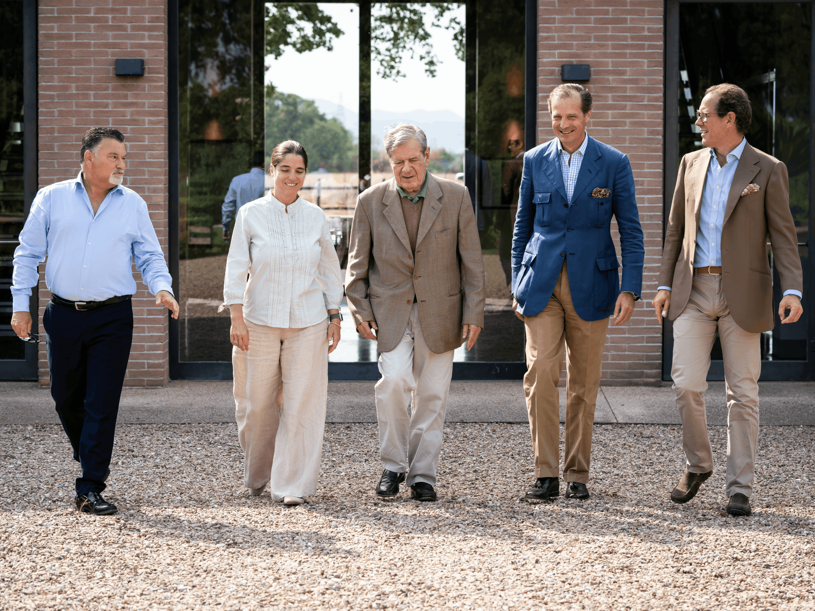 Five people in business attire walk on a gravel path, smiling. Brick building and greenery in the background, evoking a relaxed, professional mood.