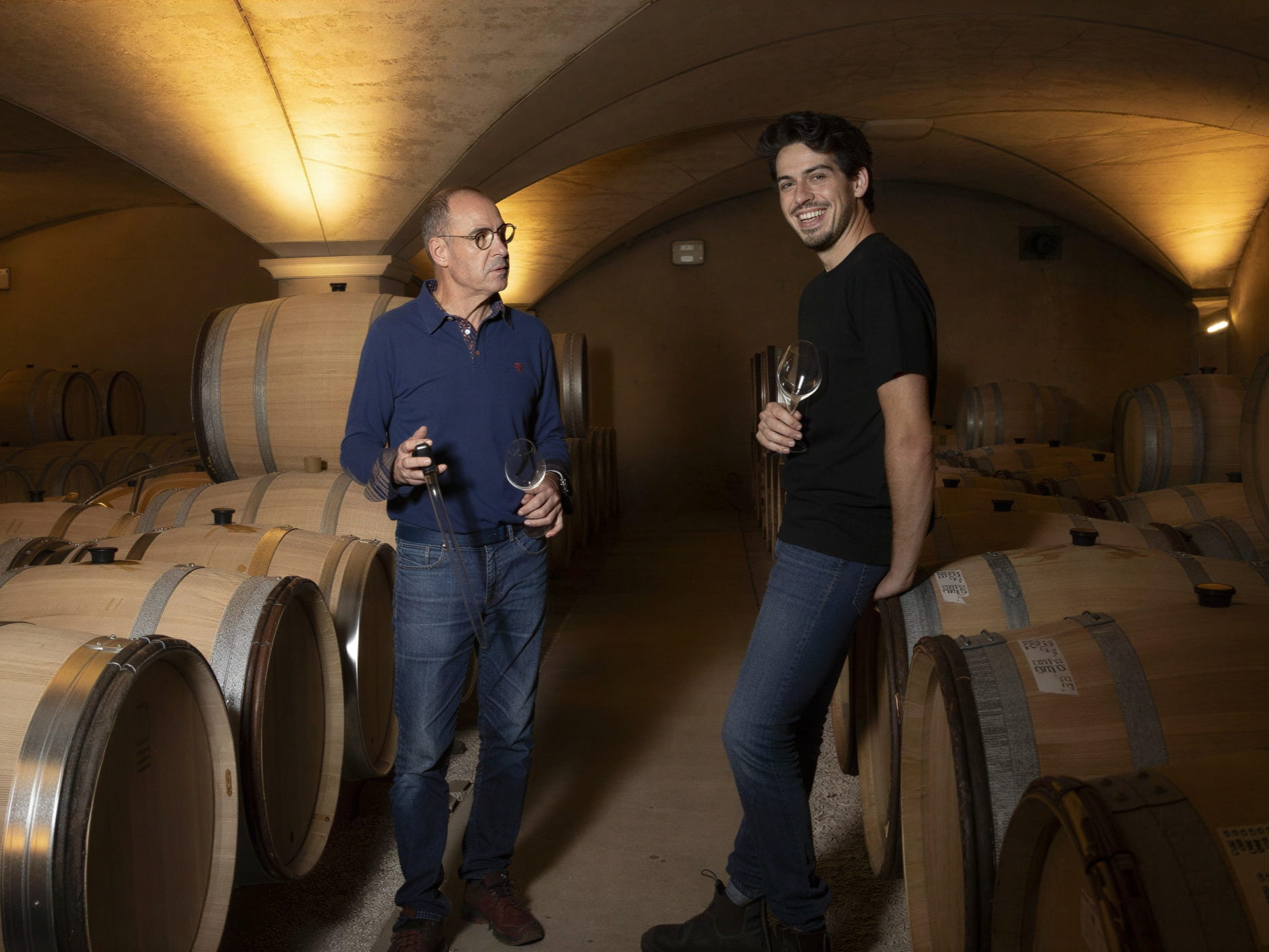 Jean Marc and Antonin Pillot in a wine cellar holding glasses, surrounded by barrels. One smiles while the other looks thoughtful. Warm lighting sets a cozy mood.