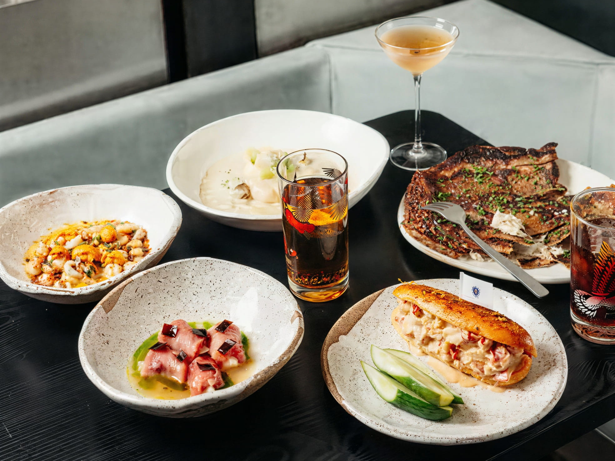 A table with a selection of dishes from Smithereens NYC including seafood, meat, and cocktails. Background has a gray sofa. Plates are speckled, and mood is elegant.