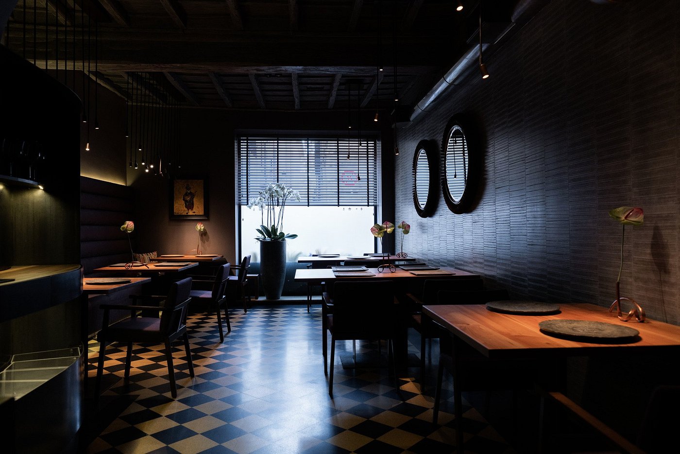 Dimly lit restaurant with wooden tables, checkered floor, and large window. Vases with flowers on tables, creating a cozy ambiance.