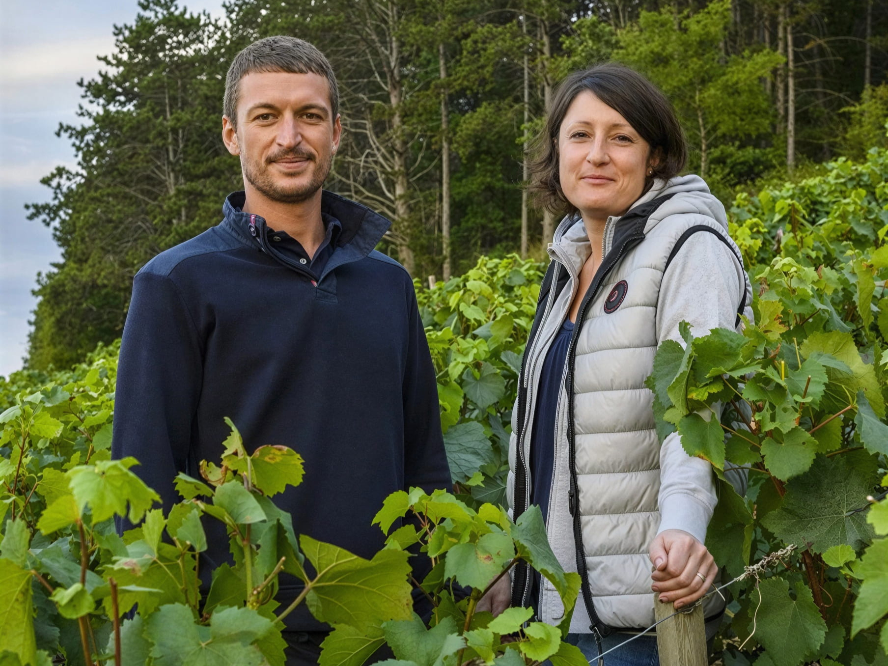 François Raveneau winemakers Isabelle and Maxime Raveneau standing in a vineyard, surrounded by green grapevines. Both are dressed casually. Trees in the background. Tranquil mood.
