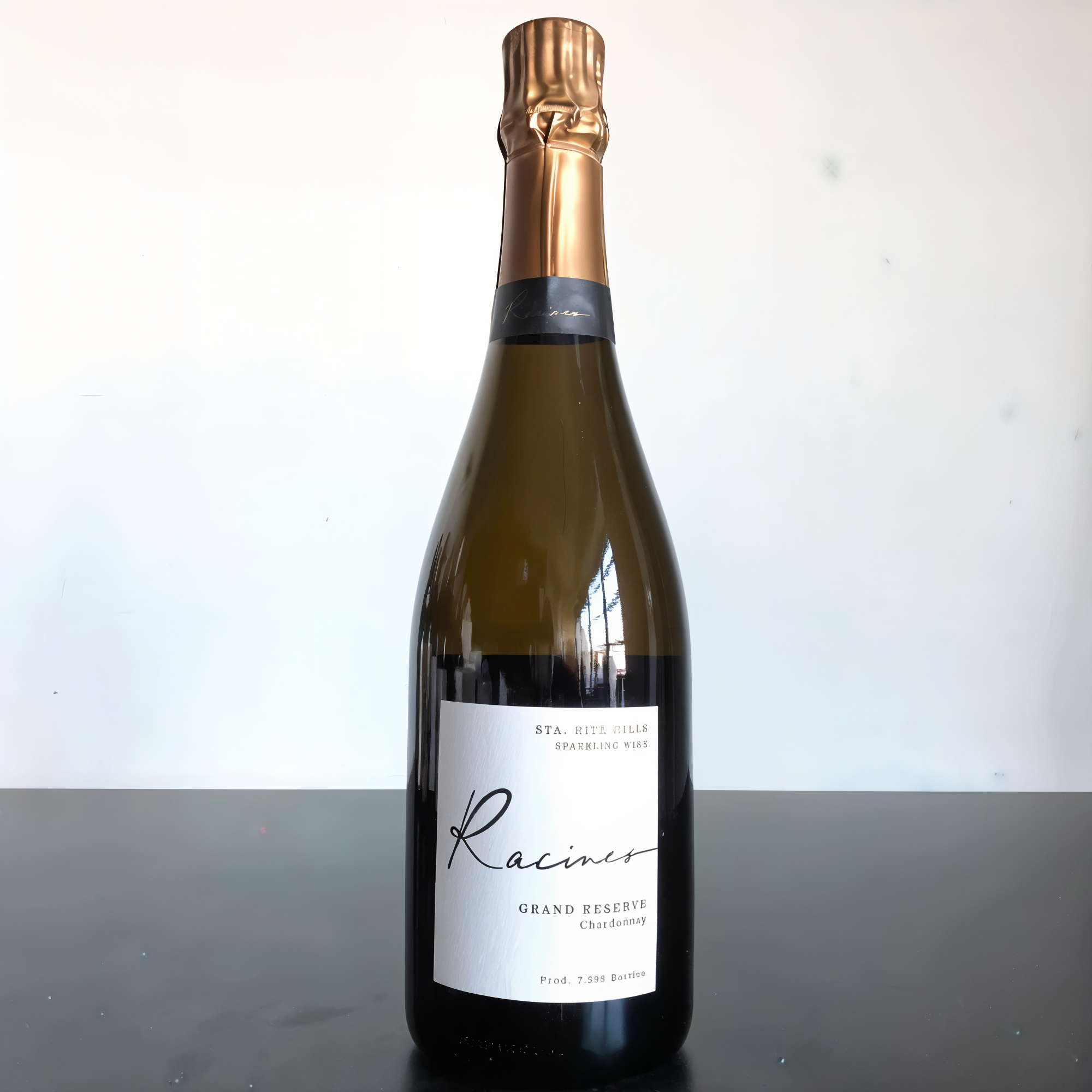 A bottle of Racines Grand Reserve Chardonnay with a gold foil top on a dark surface. Label text visible. Background is a light gradient.